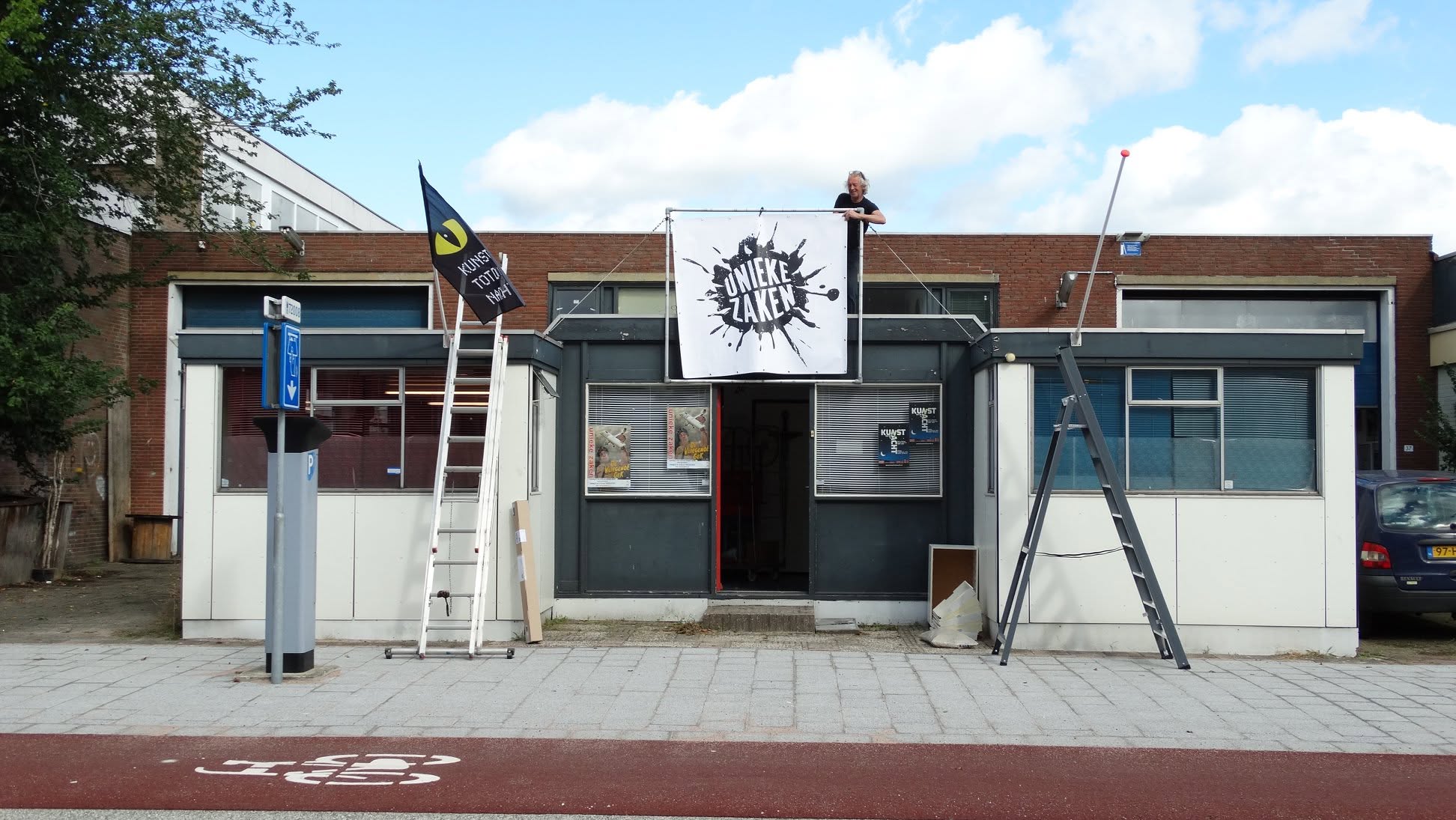 Voorkant van een gebouw met ladders en een groot spandoek met de tekst Unieke Zaken, met iemand op het dak