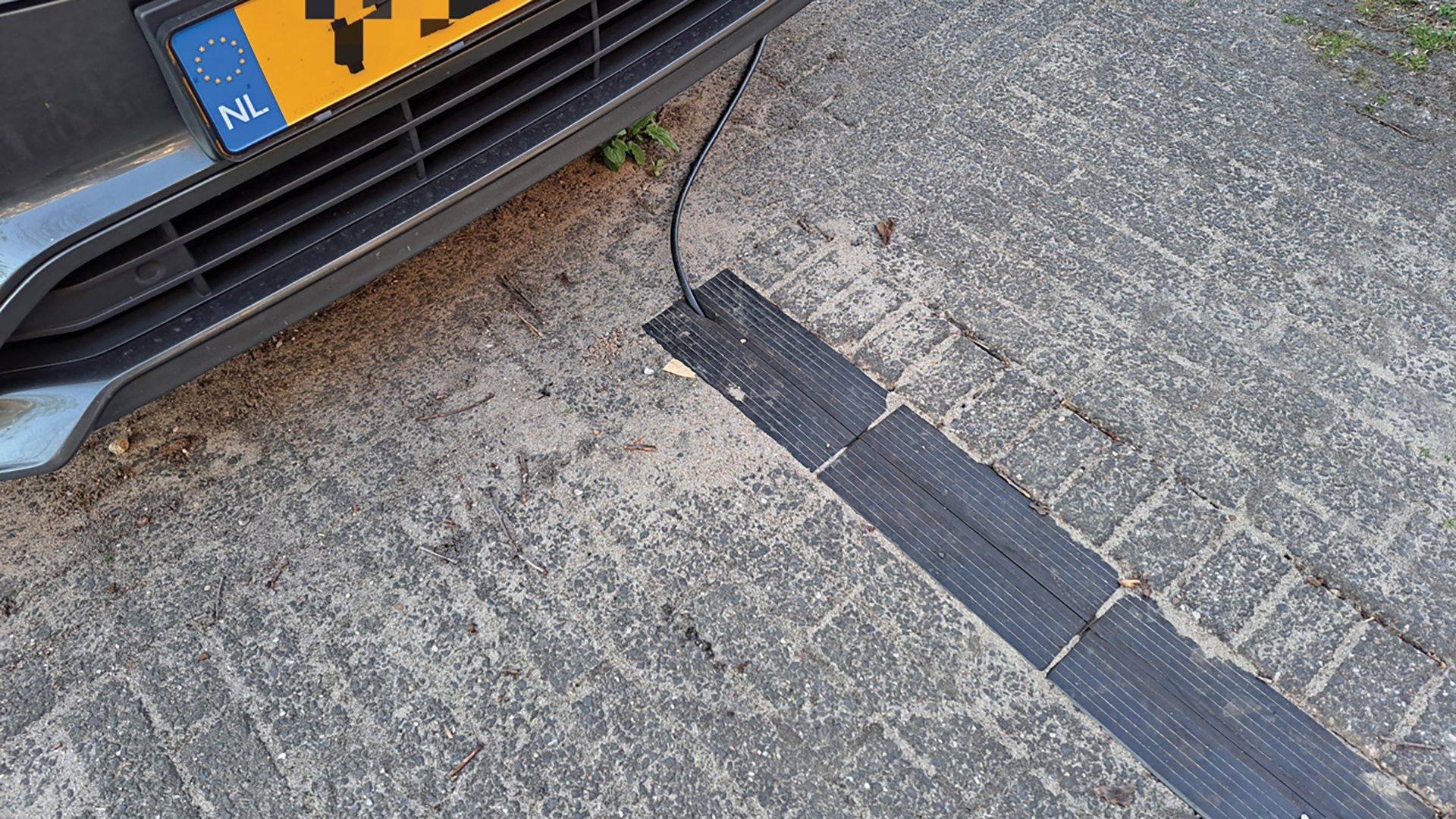 Voorkant van een auto met Nederlands kenteken; kabel loopt naar zwarte rubberen kabelgoot op de straat