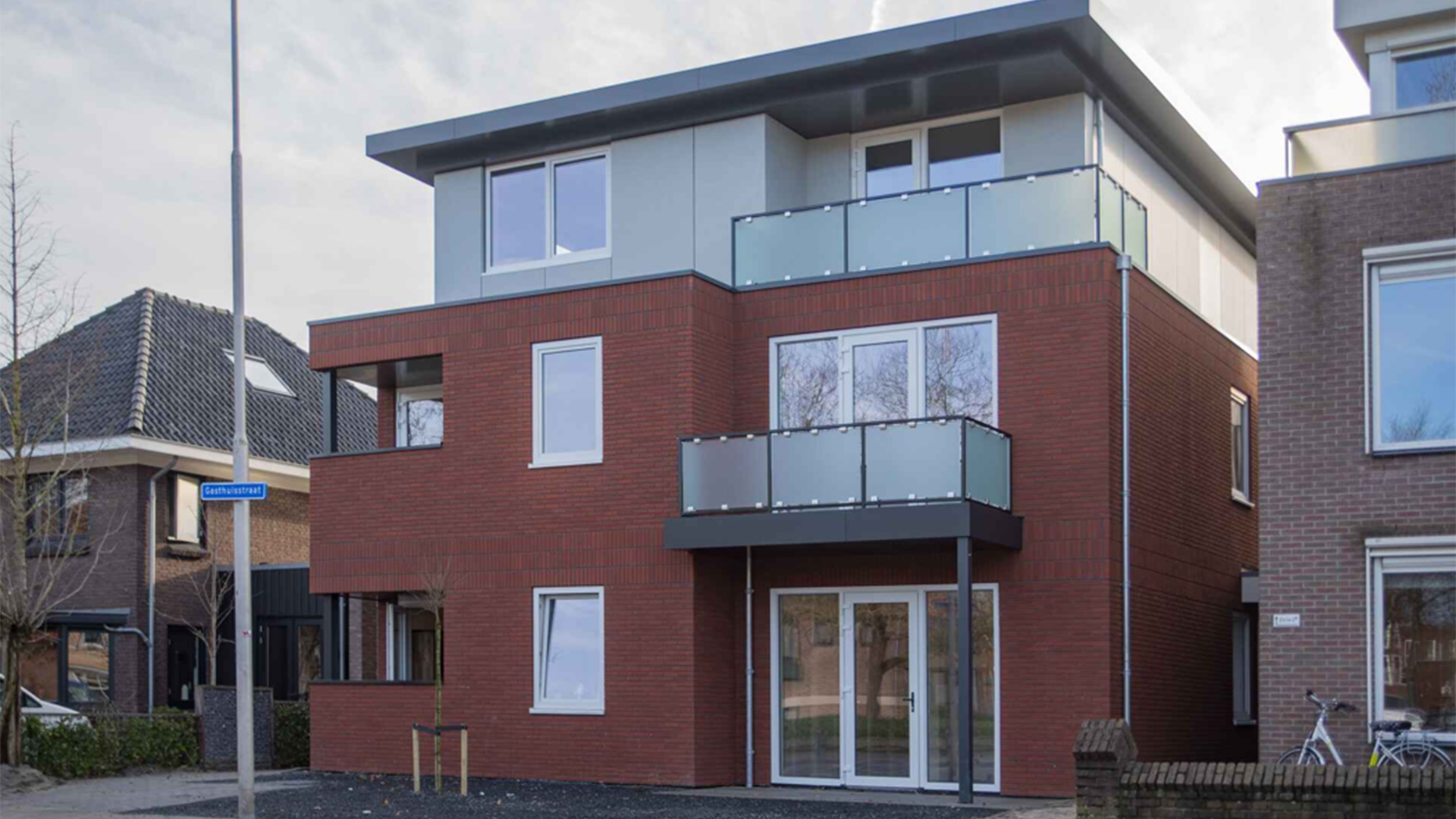 Modern roodstenen hoekwoning met twee balkons met matglas en een lichtgrijze bovenverdieping met plat dak
