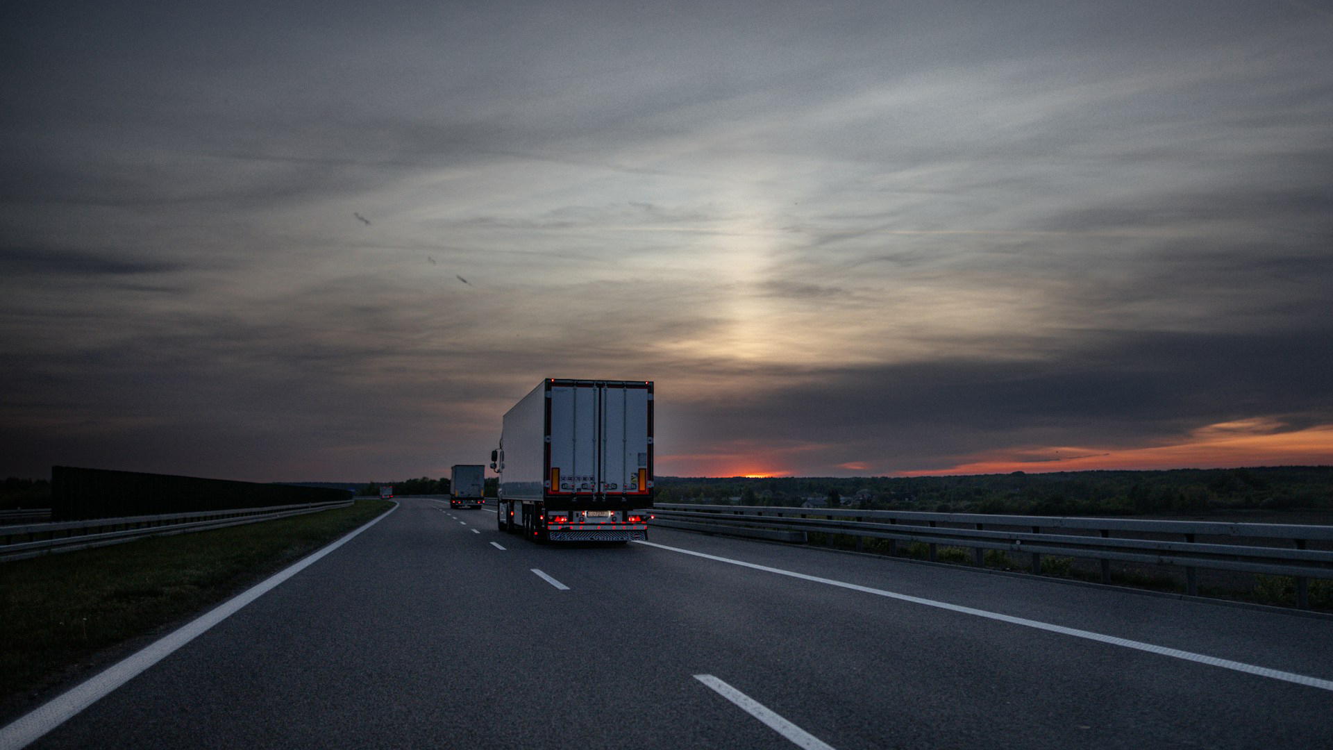 Vrachtwagen rijdt op een snelweg bij zonsondergang onder een bewolkte hemel, met nog een vrachtwagen verderop