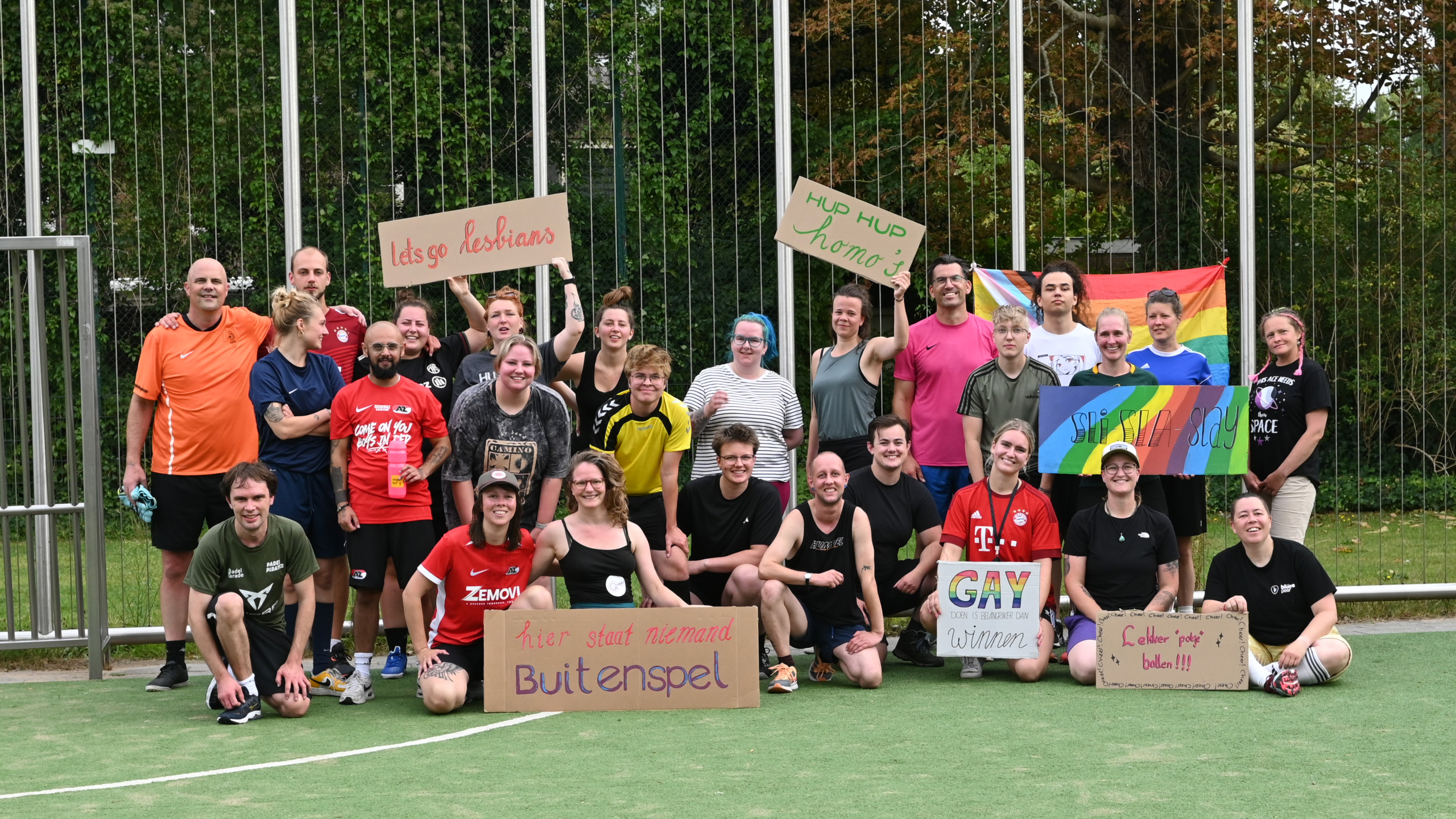Groepsfoto op sportveld met regenboogvlag en borden met LHBTQ+-teksten, waaronder “Hup hup homo’s” en “GAY”
