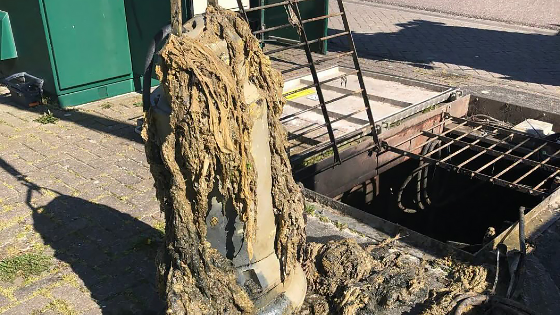 Overlopende rioolput met bruine modder en slib op trottoir naast open mangat en metalen rooster