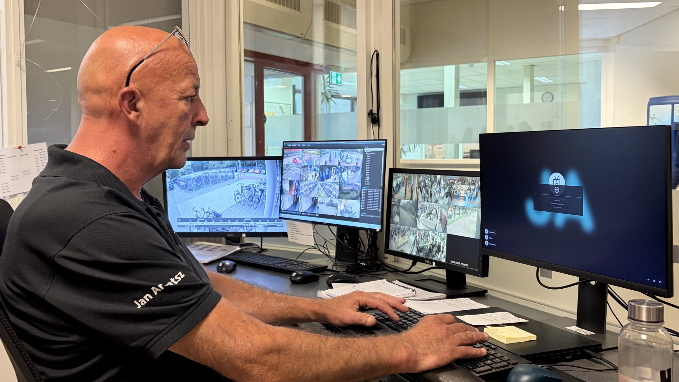 Beveiliger in controlekamer bekijkt camerabeelden op meerdere monitors en bedient toetsenbord en muis