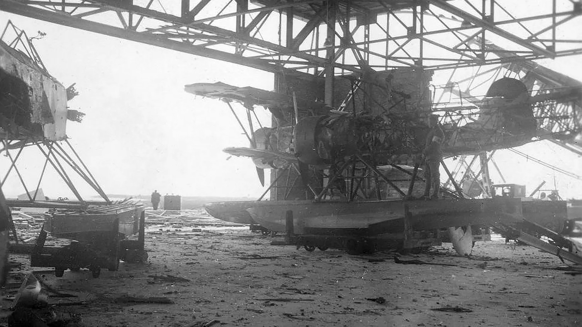 Zwart-witfoto van een beschadigd watervliegtuig tussen metalen draagconstructies in een hangar op een verlaten terrein