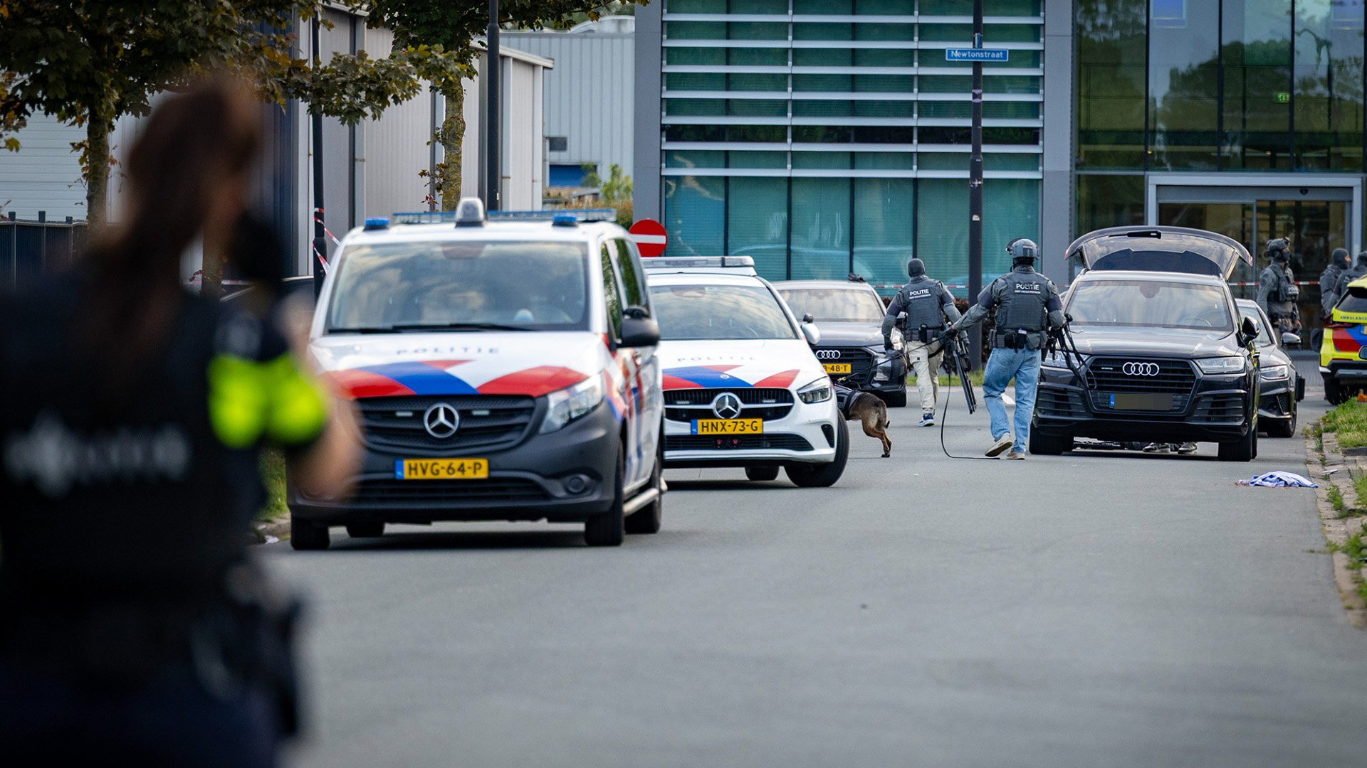 Politie met kogelwerende vesten en hond bij politiewagens op straat, met een auto met open achterklep