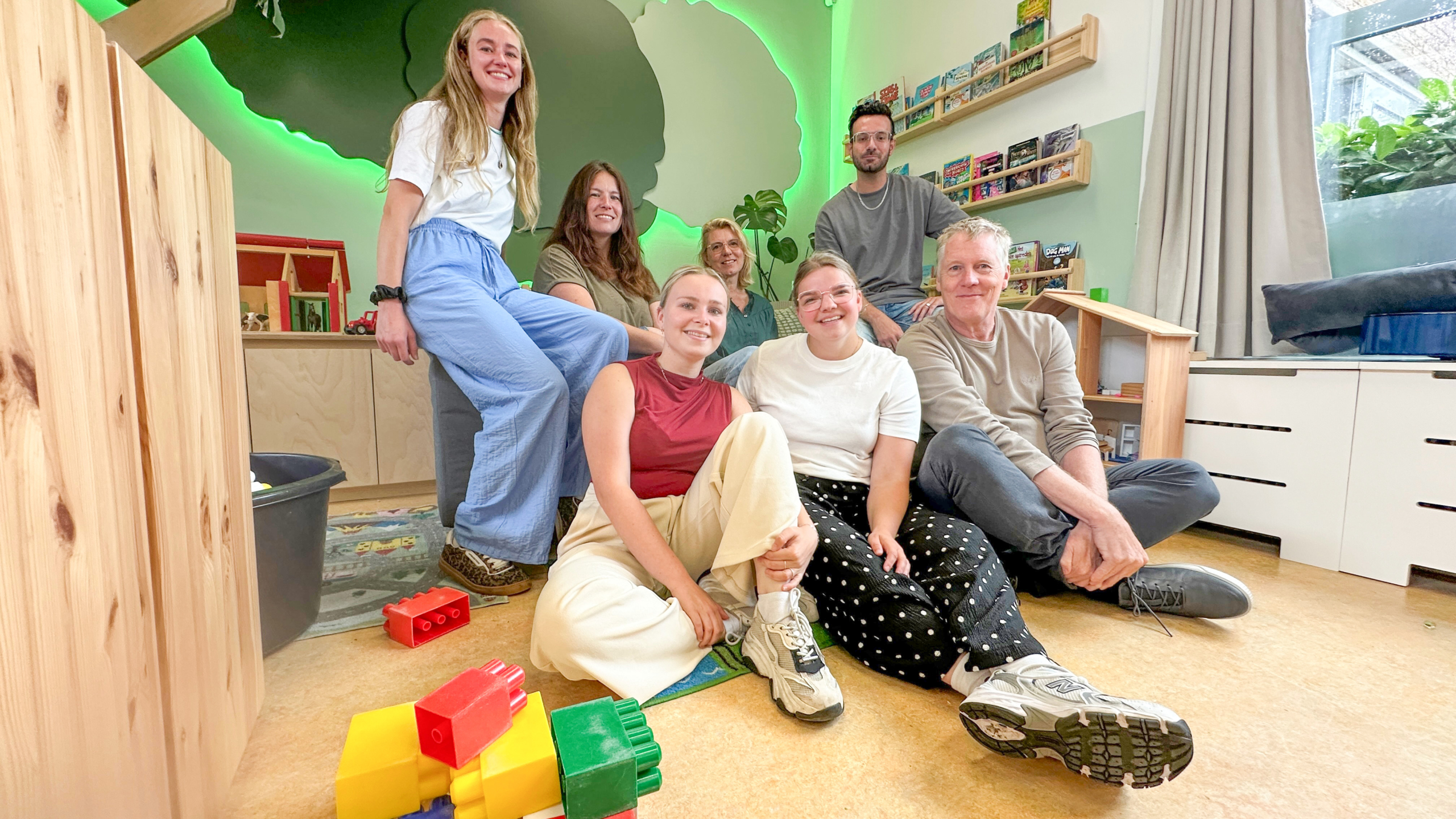 Groep van zes mensen poseert glimlachend in een kinderruimte met speelgoedblokken, boekenplanken en groene wandverlichting