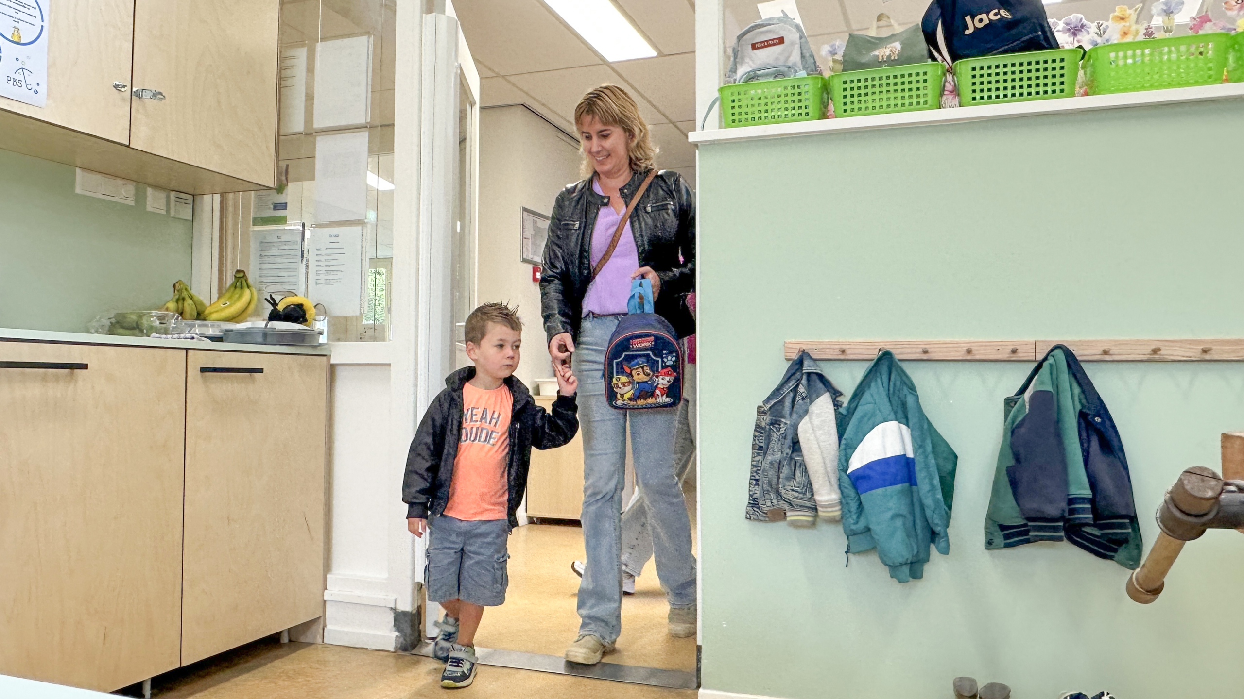 Vrouw met rugzak brengt jongen aan de hand een klaslokaal binnen, met jassen aan kapstok en mandjes op plank