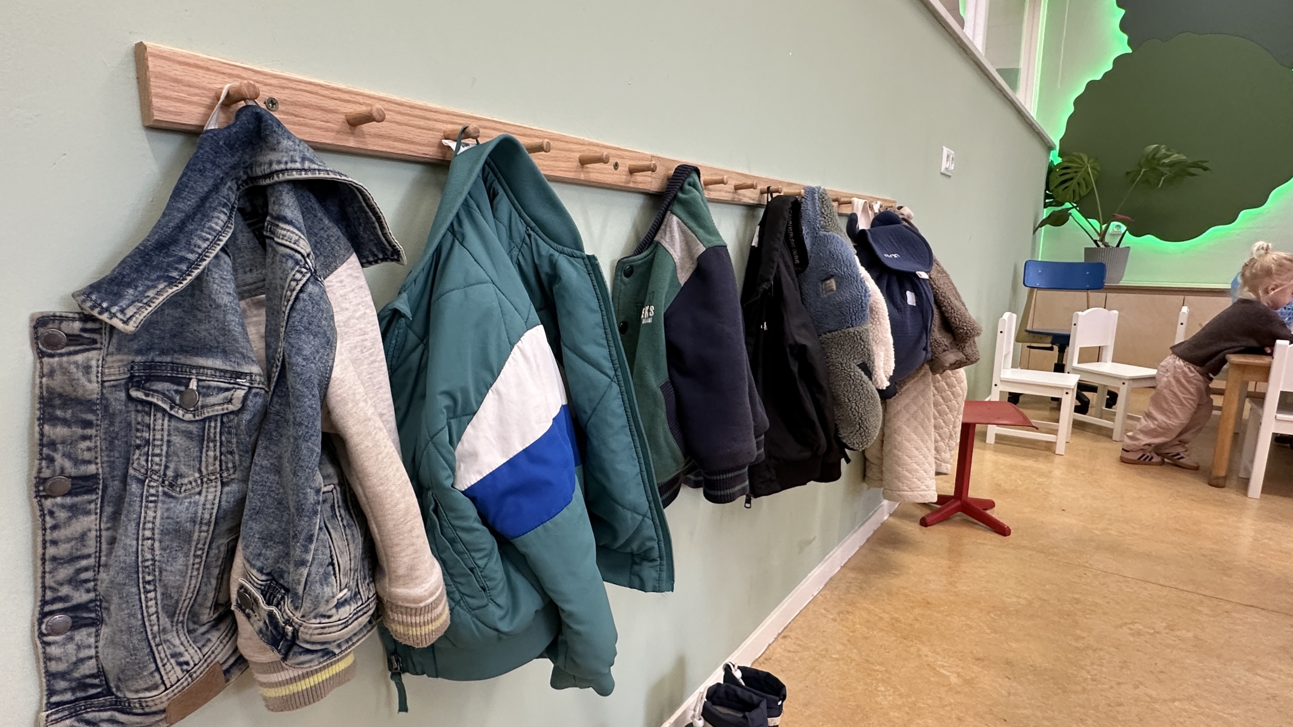 Jassen en kleding hangen aan een houten kapstok in een kinderdagverblijf; op de achtergrond staan tafels en stoeltjes