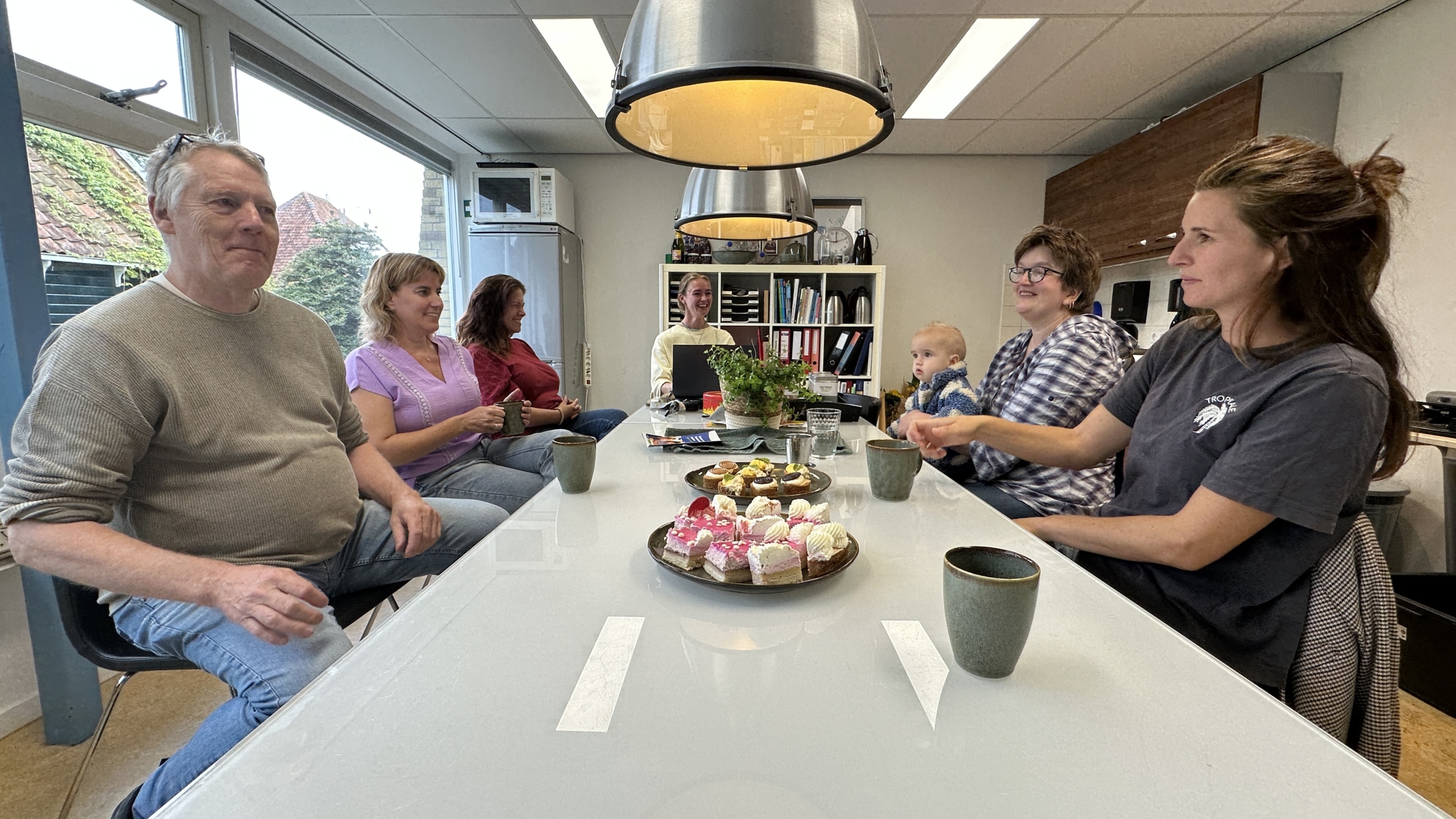 Groep collega’s zit rond een tafel in de kantine met koffie en gebak, terwijl een baby op schoot meekijkt