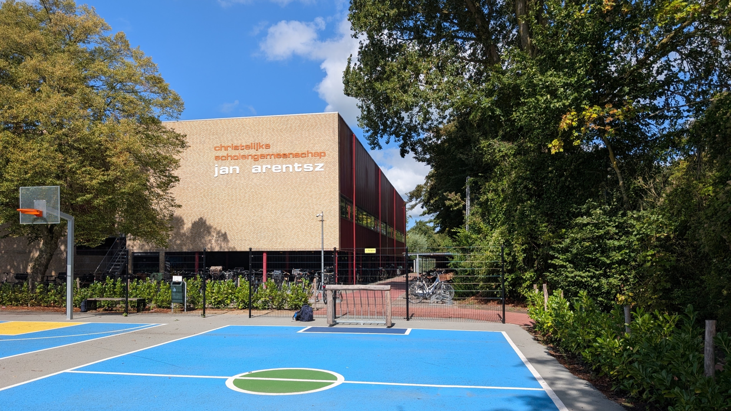 Schoolgebouw met opschrift christelijke scholengemeenschap Jan Arentsz, naast basketbalveld en fietsenrekken tussen bomen