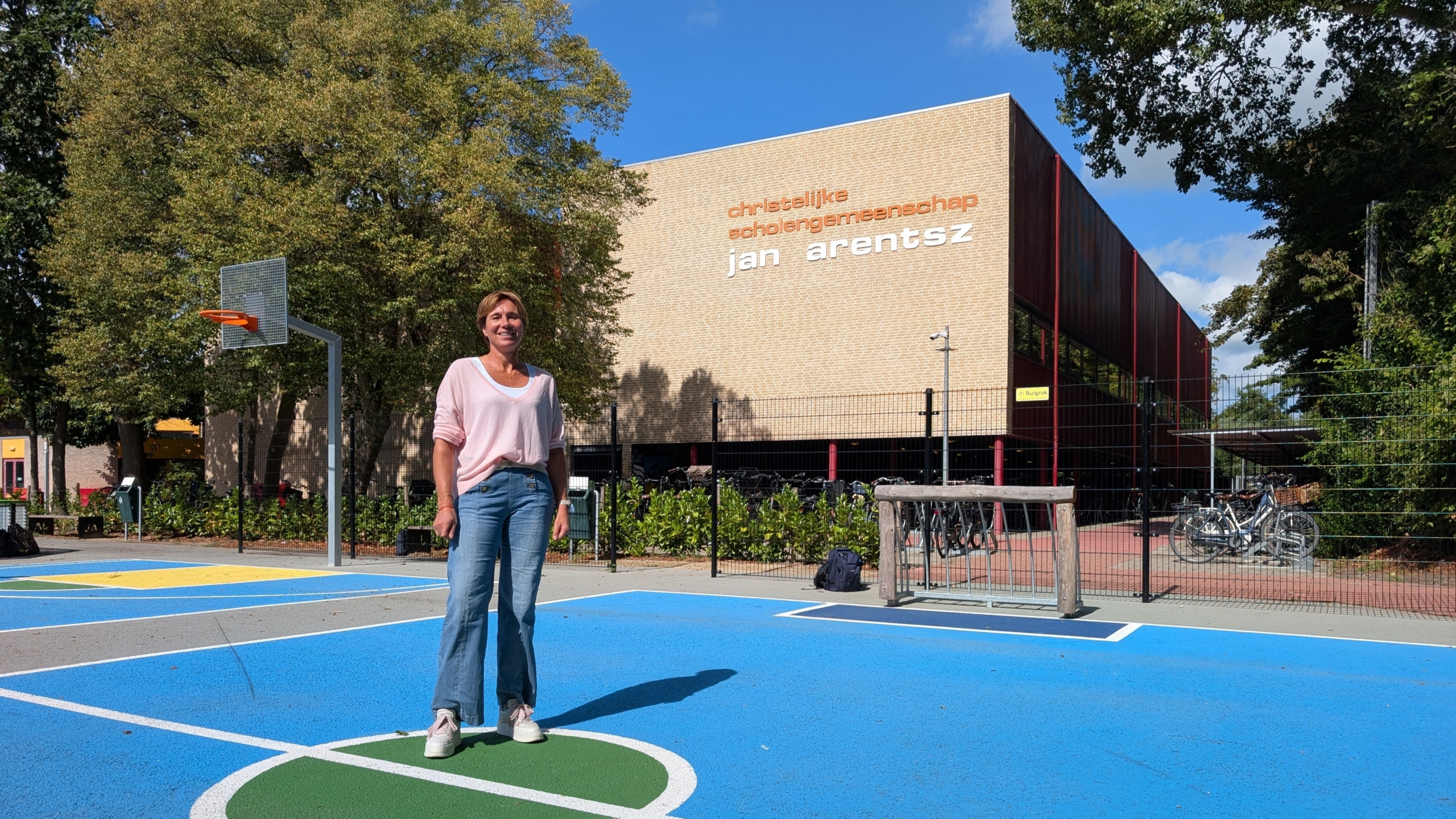 Vrouw op een blauw schoolplein met basketbalpaal voor het gebouw van christelijke scholengemeenschap Jan Arentsz