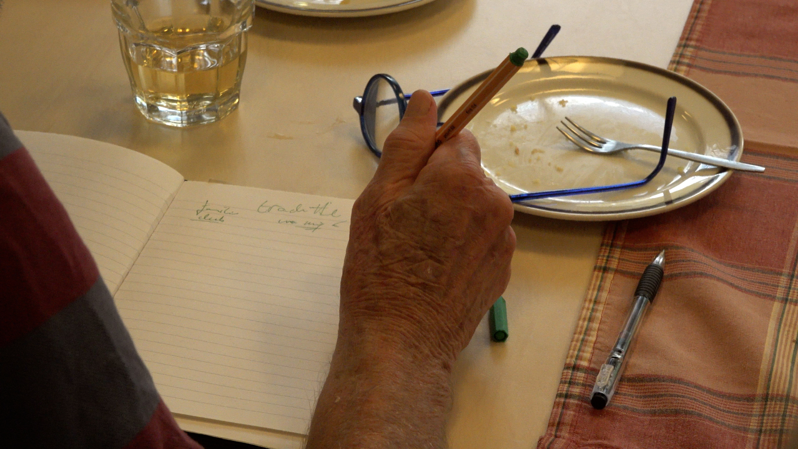 Hand houdt een potlood boven een notitieboek op tafel, met bril, leeg bord met vork en een glas erbij