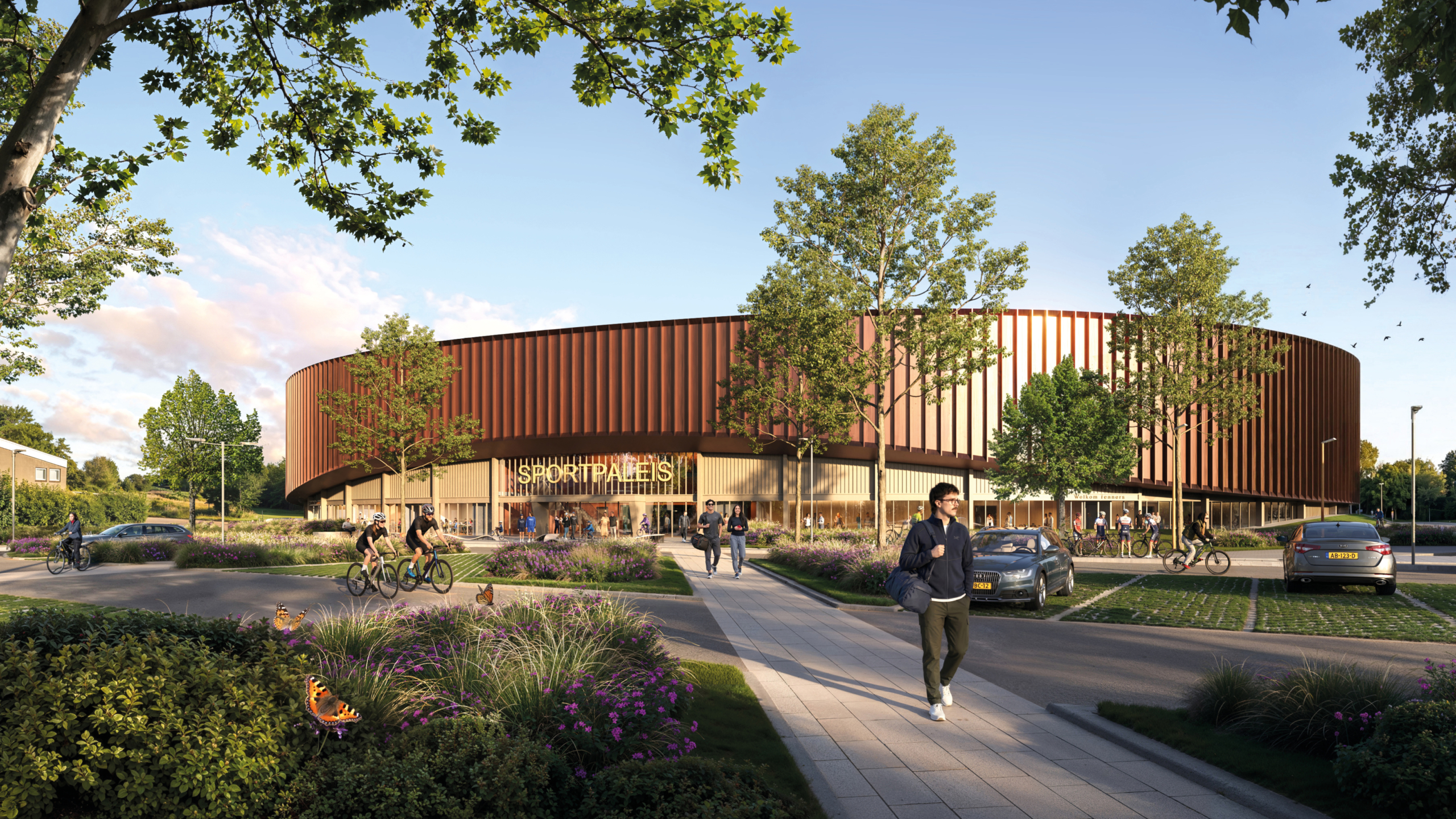 Modern rond sportpaleis met roodbruine gevel, mensen en fietsers bij de entree, omringd door groen en bloemen