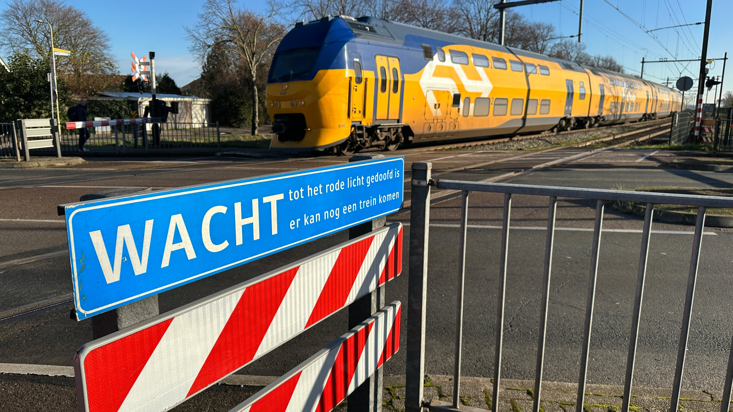 NS-dubbeldekker rijdt langs een overweg, met op de voorgrond bord: WACHT tot het rode licht gedoofd is