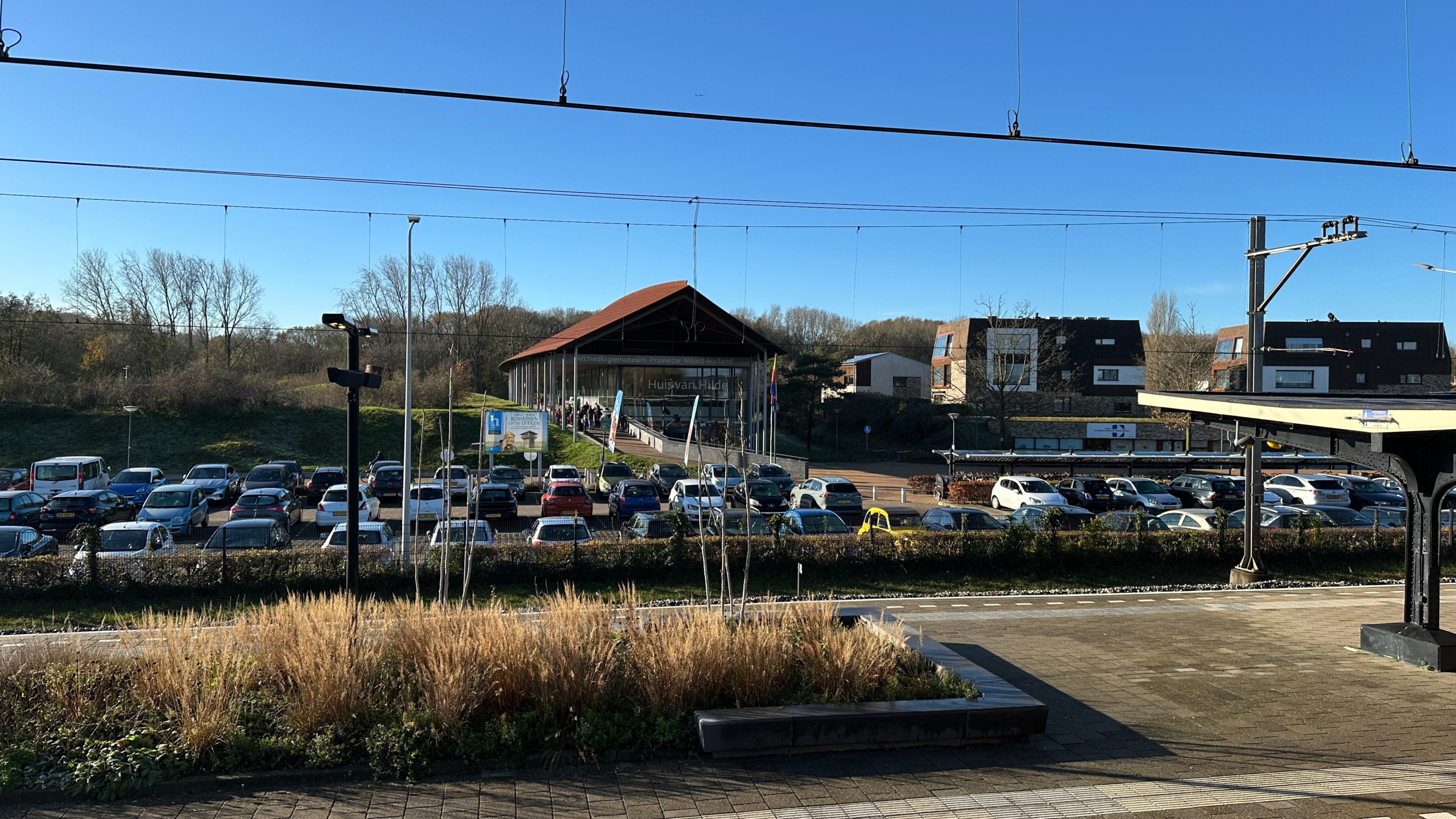 Parkeerterrein met auto’s voor het gebouw van archeologiemuseum Huis van Hilde, gezien vanaf een station perron