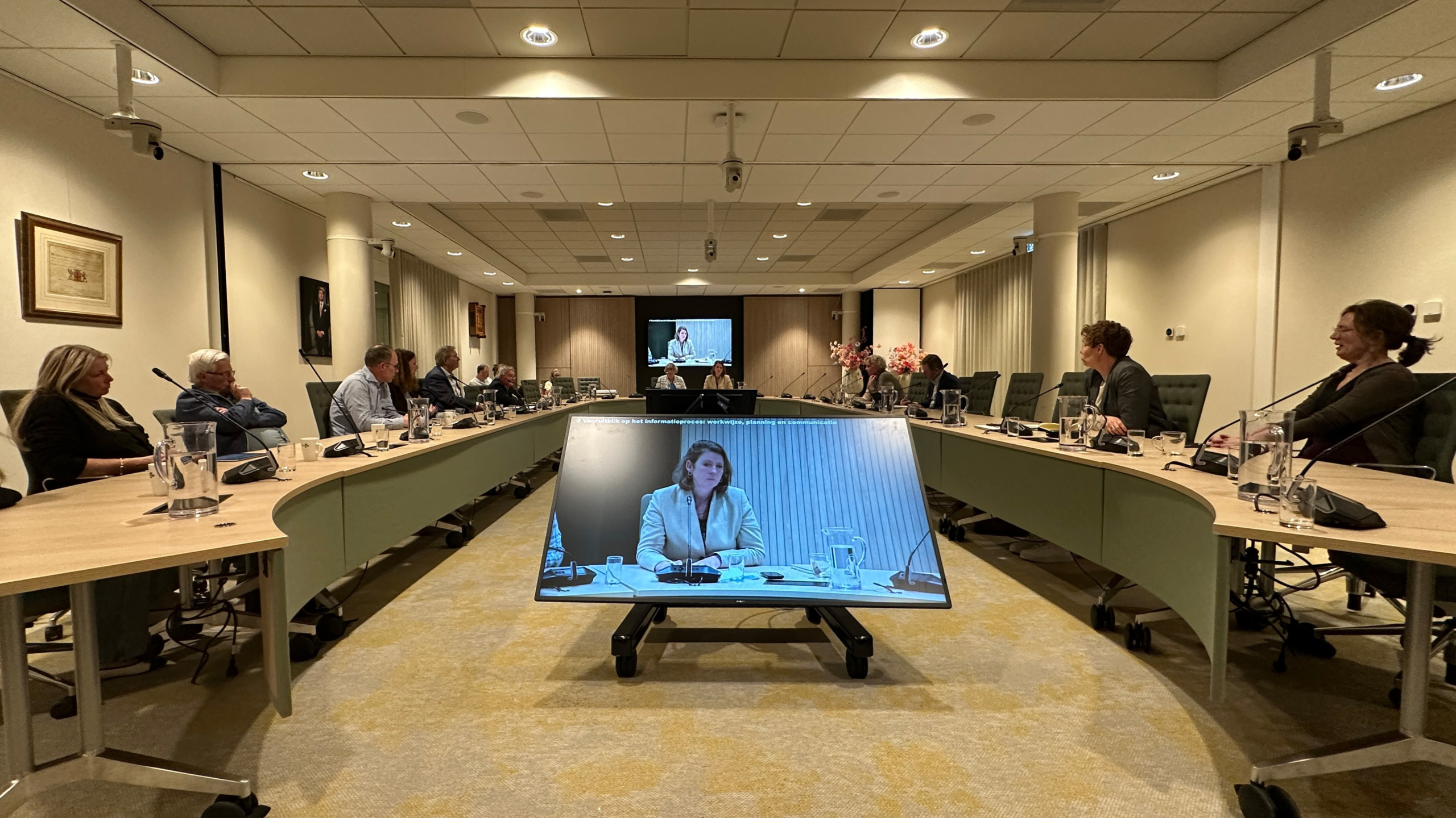 Vergaderzaal met mensen rond een U-vormige tafel, kijkend naar een groot scherm met een spreker via videoverbinding