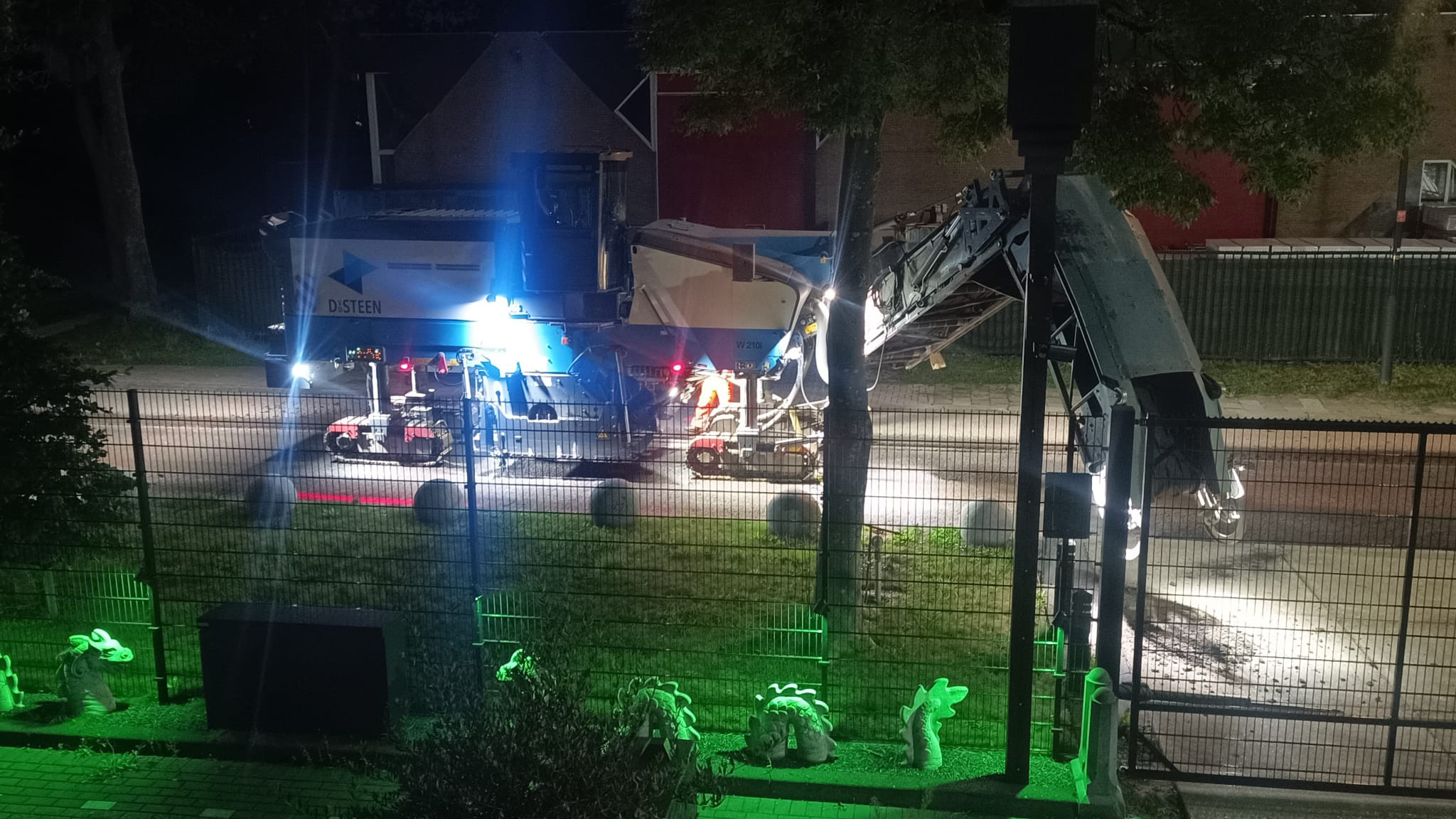 Wegfreesmachine met transportband werkt ’s nachts op straat, fel verlicht achter een hek