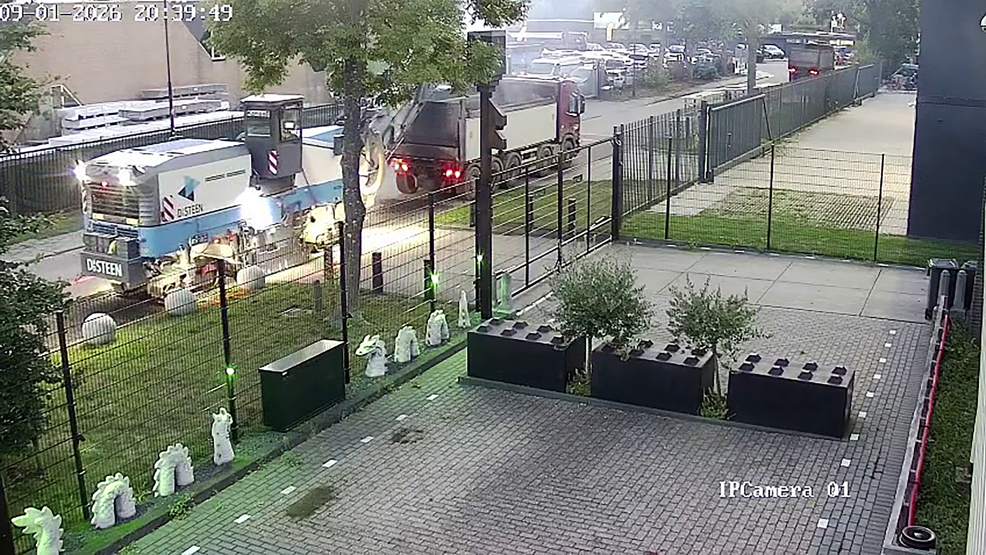 Bewakingscamerabeeld van een wegmachine en vrachtwagen achter een hek langs een weg, met bestrate binnenplaats op de voorgrond