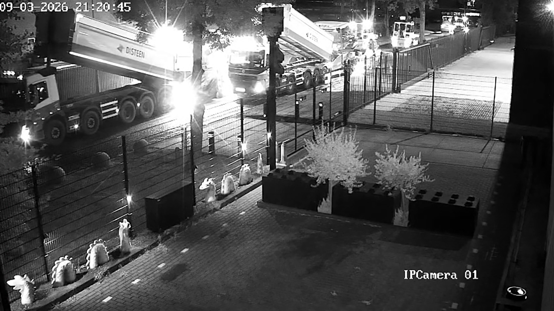 Nachtelijke CCTV-opname van twee kiepwagens met lichten aan op een weg naast een hek en parkeerterrein