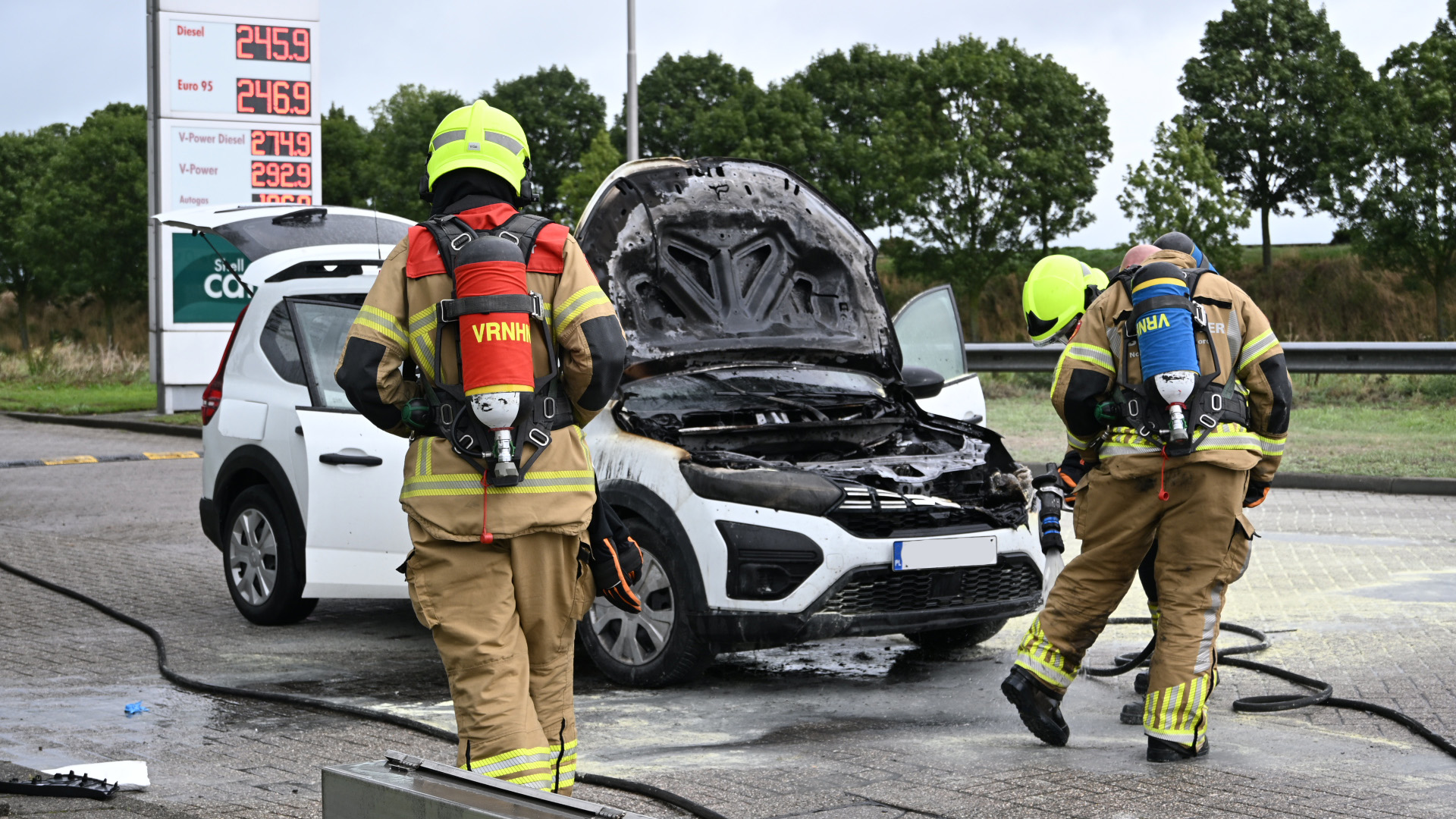 Brandweerlieden blussen smeulende auto met open motorkap bij tankstation; voorkant zwaar zwartgeblakerd