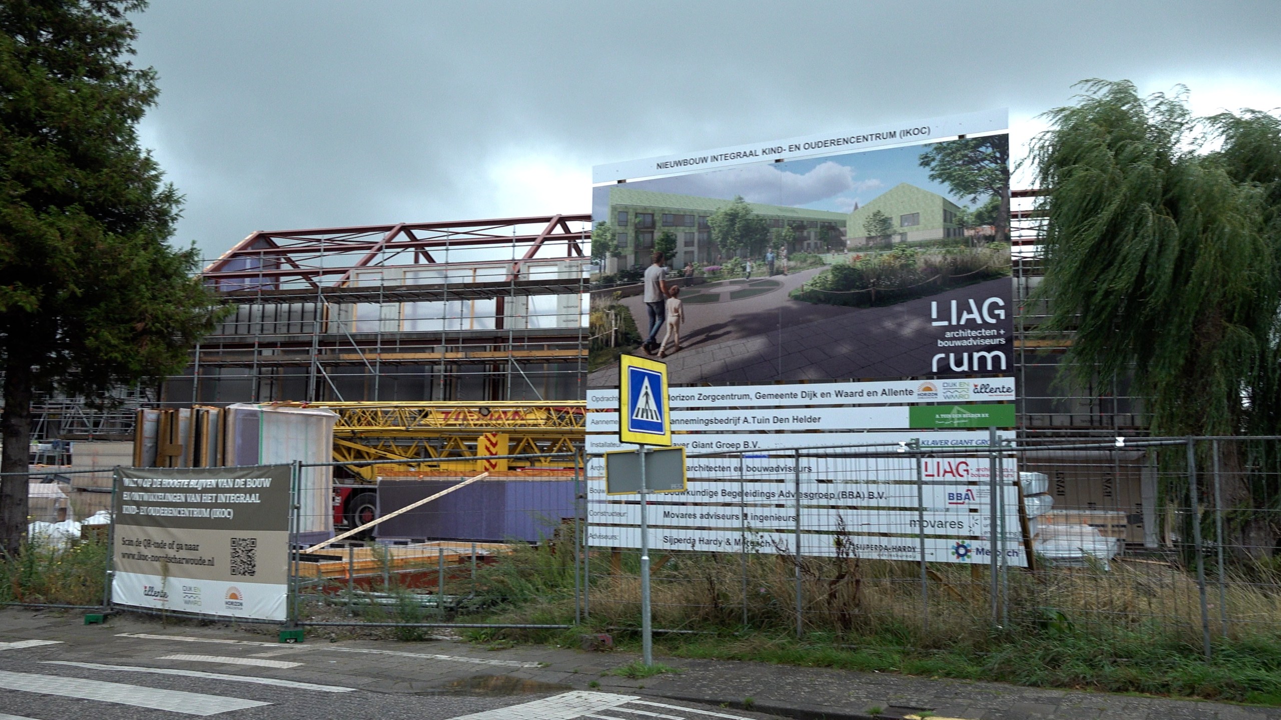 Bouwplaats met steigers en kraan, met groot bord van nieuwbouw kind- en ouderencentrum (IKOC)