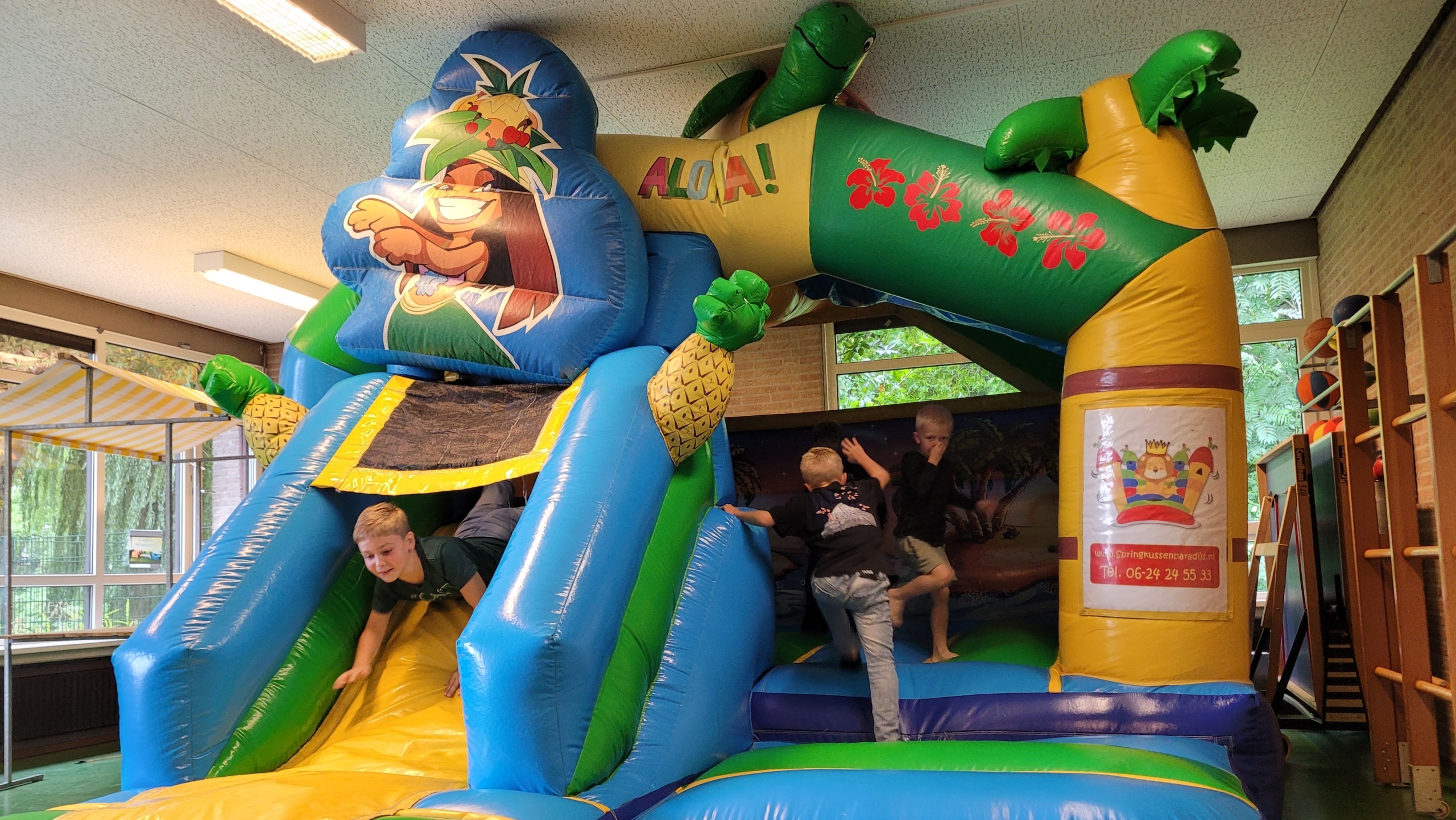 Kinderen spelen op een grote opblaasbare springkussen-glijbaan met tropisch thema en palmboomboog met tekst ALOHA