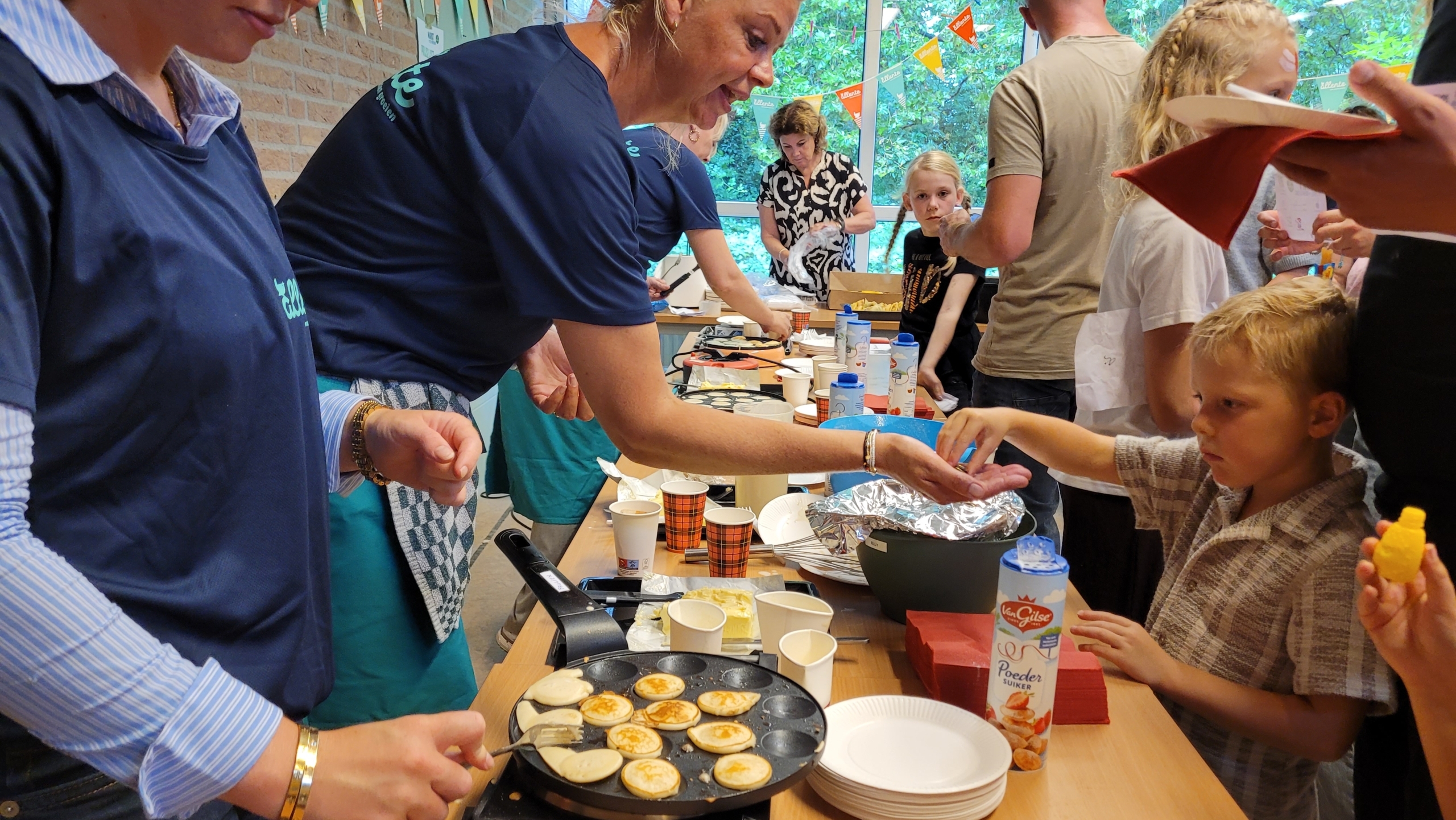 Vrijwilligers bakken poffertjes en serveren ze aan kinderen aan een drukke tafel met borden, bekers en poedersuiker