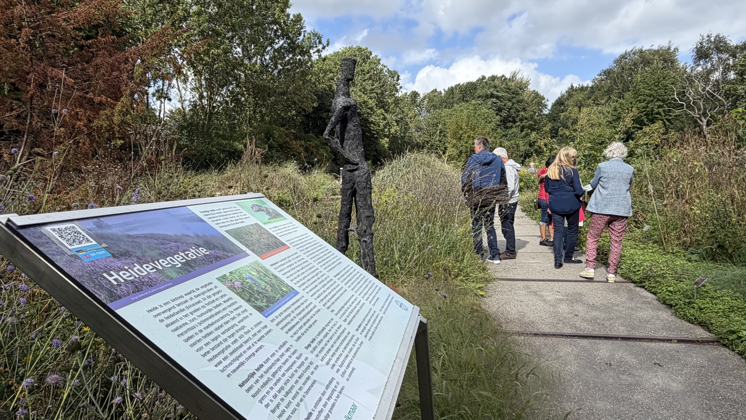 Informatiepaneel met titel Heidevegetatie langs een tuinpad, met bezoekers en een donkere mensfiguur-sculptuur tussen het groen