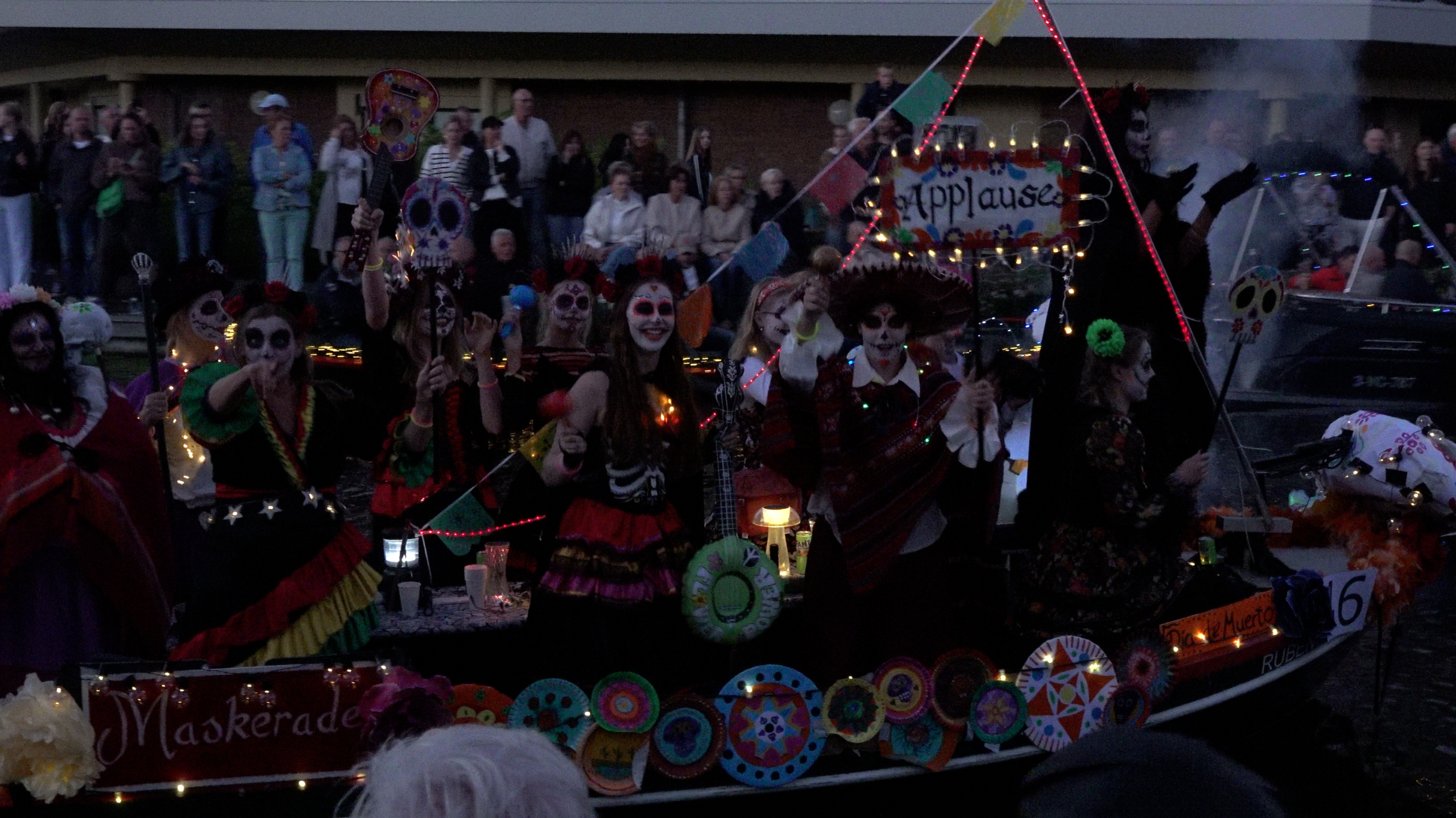 Groep verklede mensen met Día de los Muertos-schmink op een verlichte praalwagen, met publiek op de achtergrond