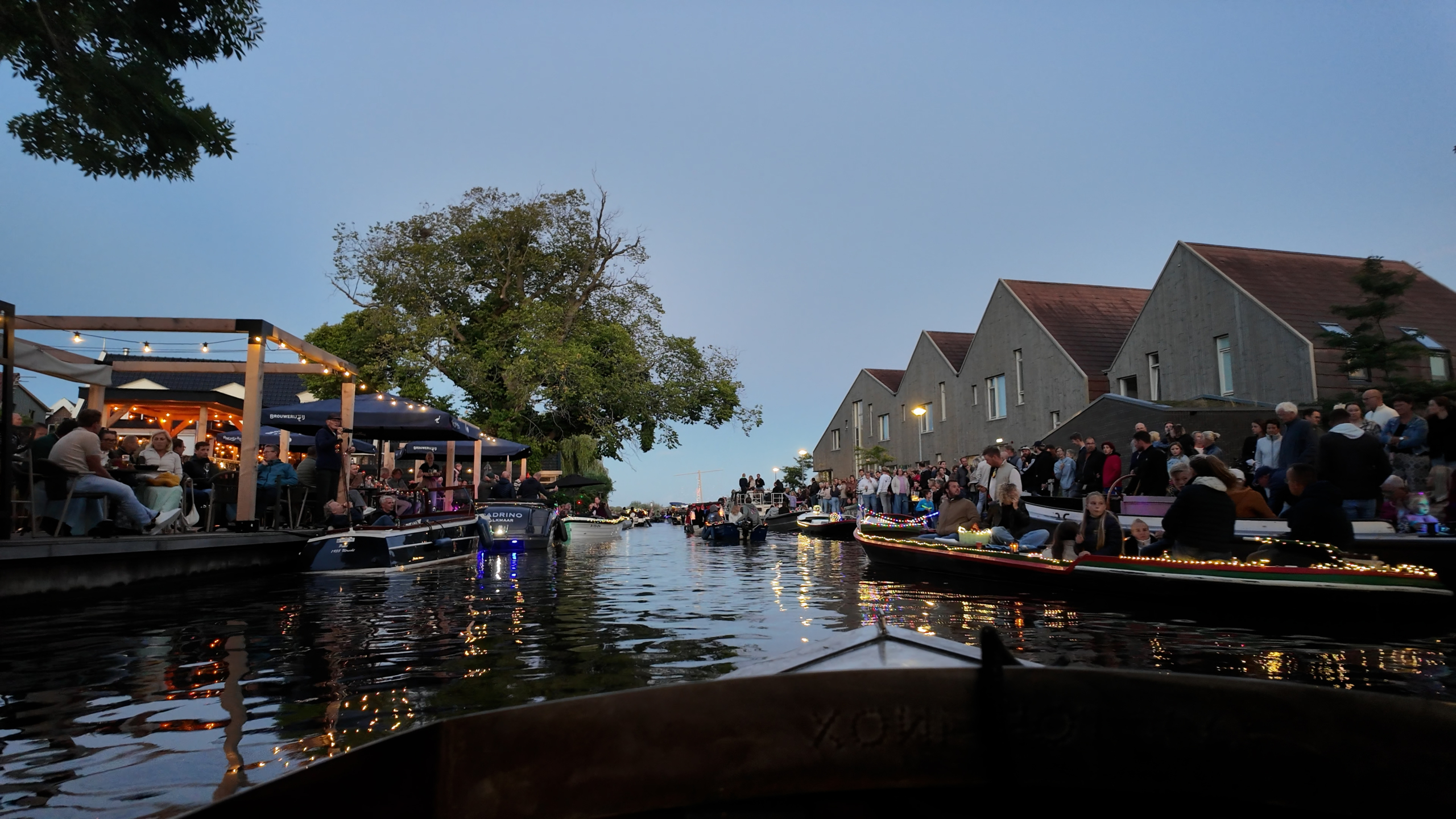 Avondbeeld van een kanaal met verlichte boten, publiek langs de kade en moderne huizen op de achtergrond
