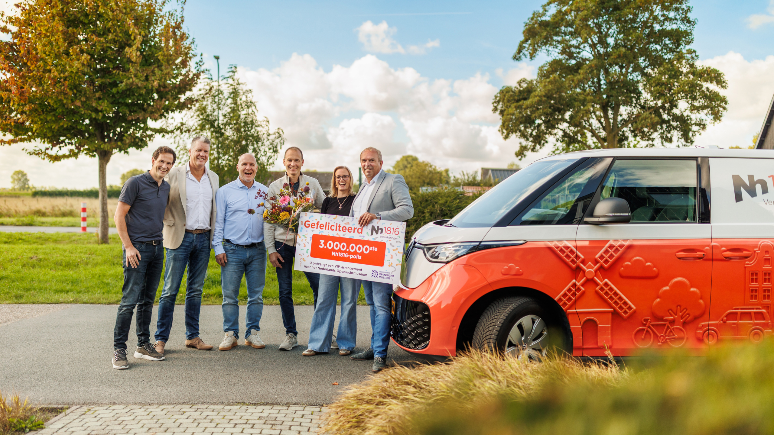 Groep mensen poseert buiten met bloemen en bord ‘Gefeliciteerd 3.000.000ste Nh1816-polis’ naast oranje busje