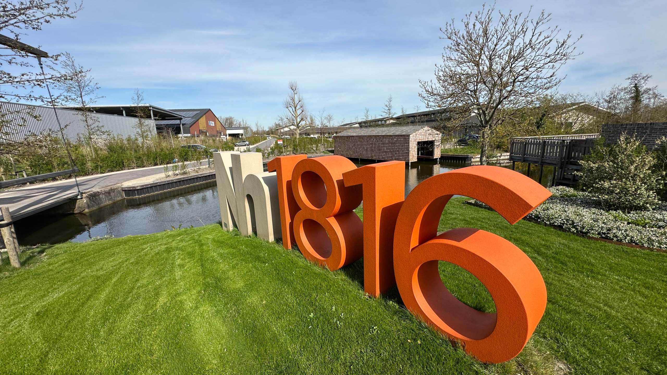 Grote sculptuur met “Nh1816” op een grasveld naast een waterpartij, met bruggetje en gebouwen op de achtergrond