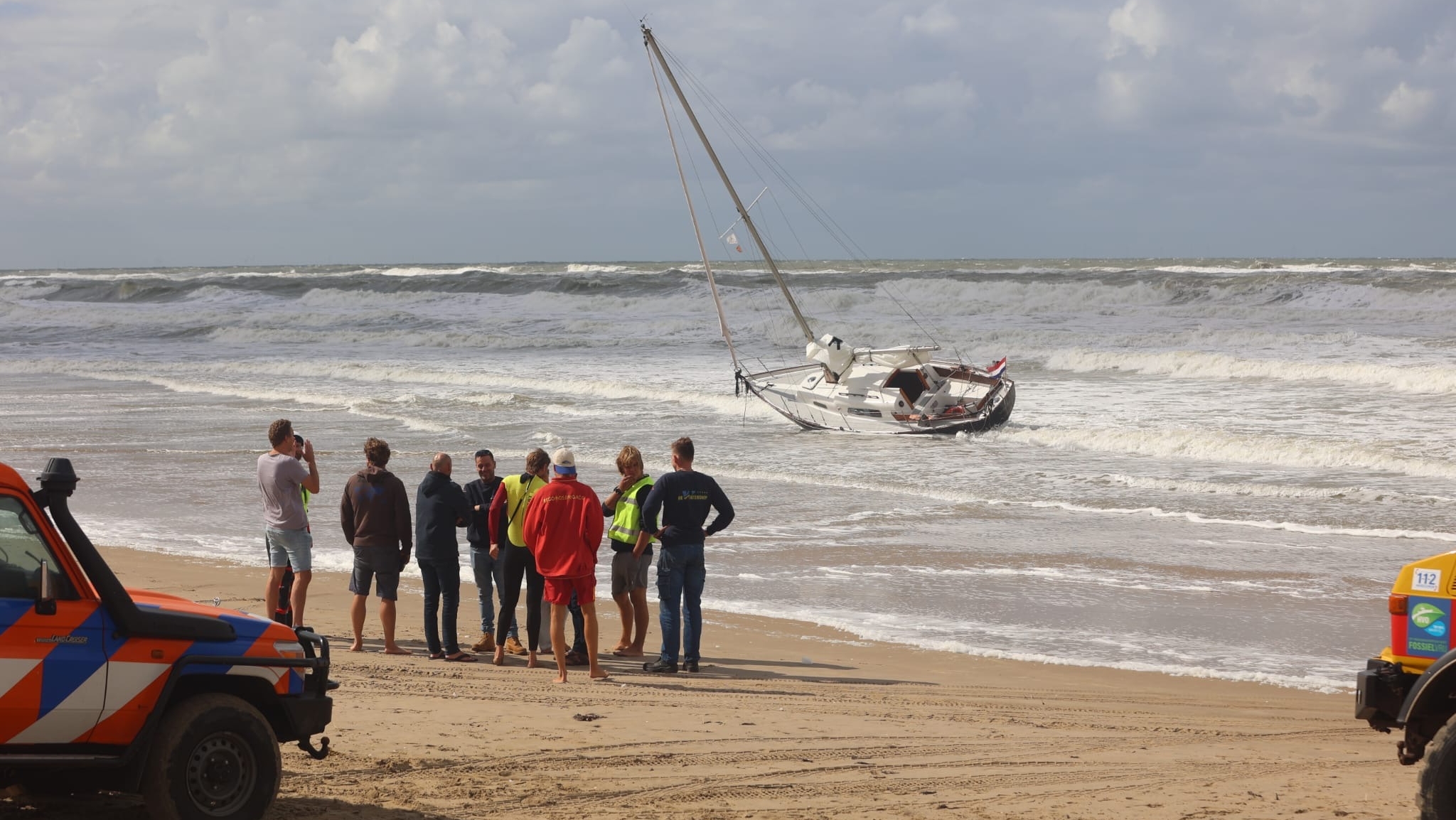 Groep hulpverleners op strand kijkt naar gekantelde zeilboot die is vastgelopen in de branding