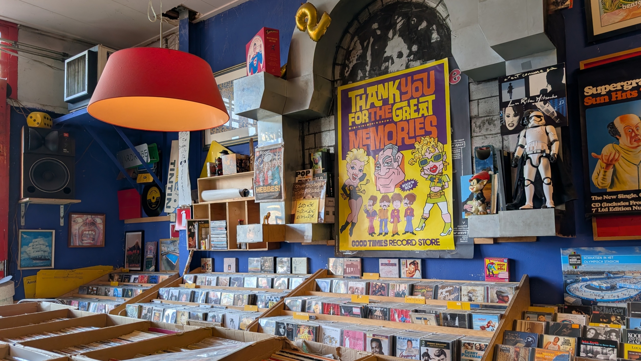 Interieur van een platenzaak met bakken vol cd’s, rode hanglamp en posters, waaronder “Thank you for the great memories”