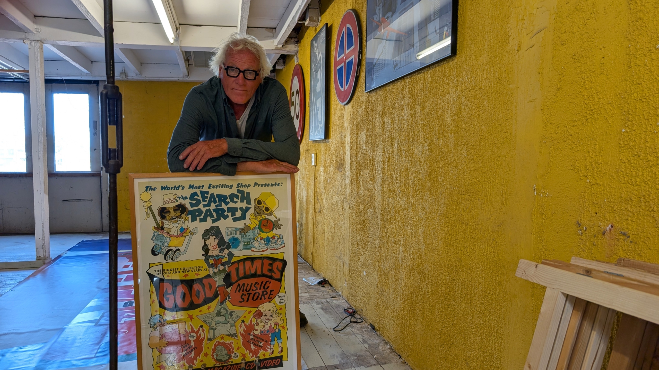 Man met grijs haar en bril leunt op een ingelijste poster in een ruimte met gele muur en verkeersborden