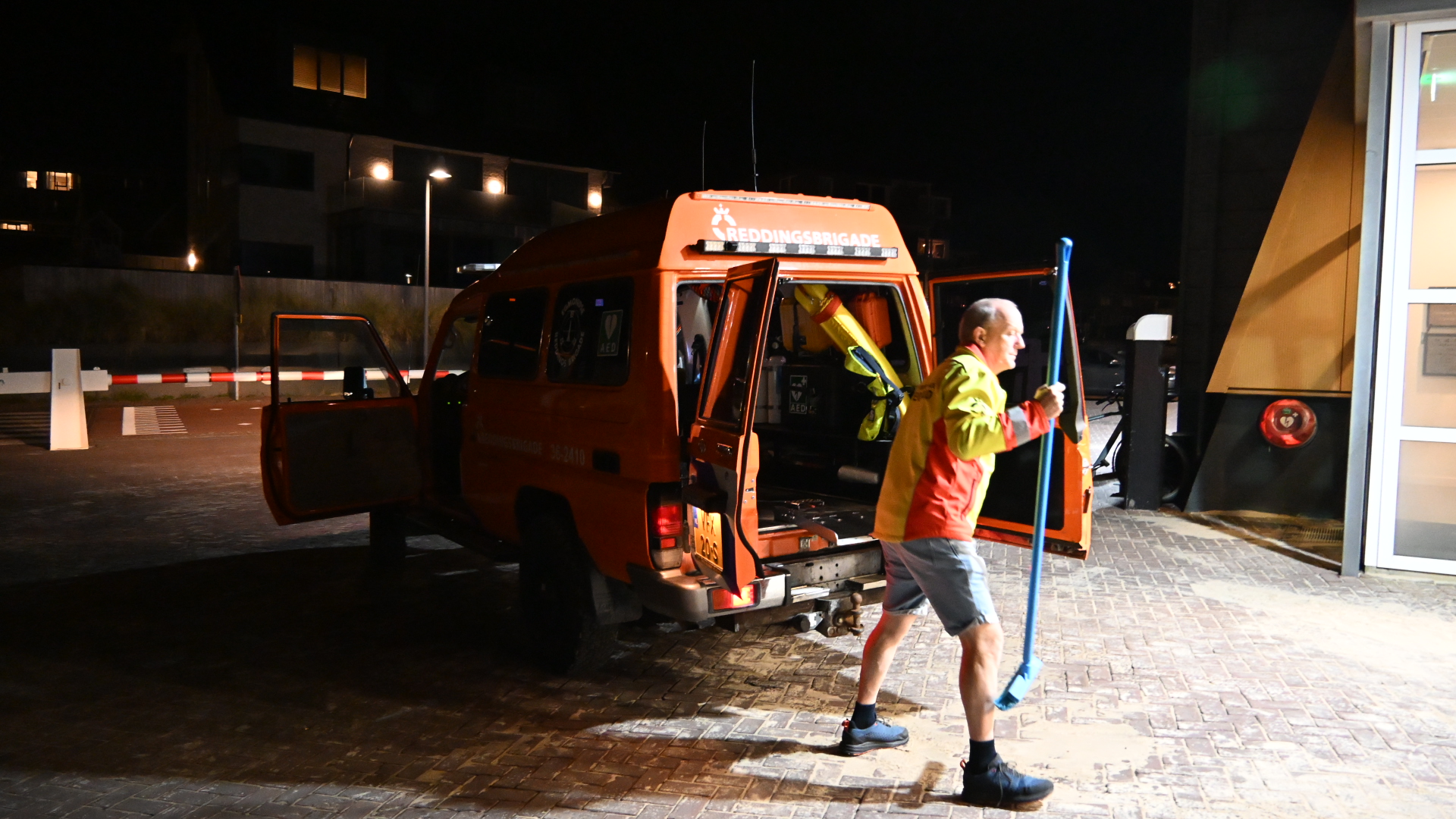 Man in rood-gele jas loopt met lange blauwe peddel naast oranje reddingsbrigadebusje met open deuren in het donker