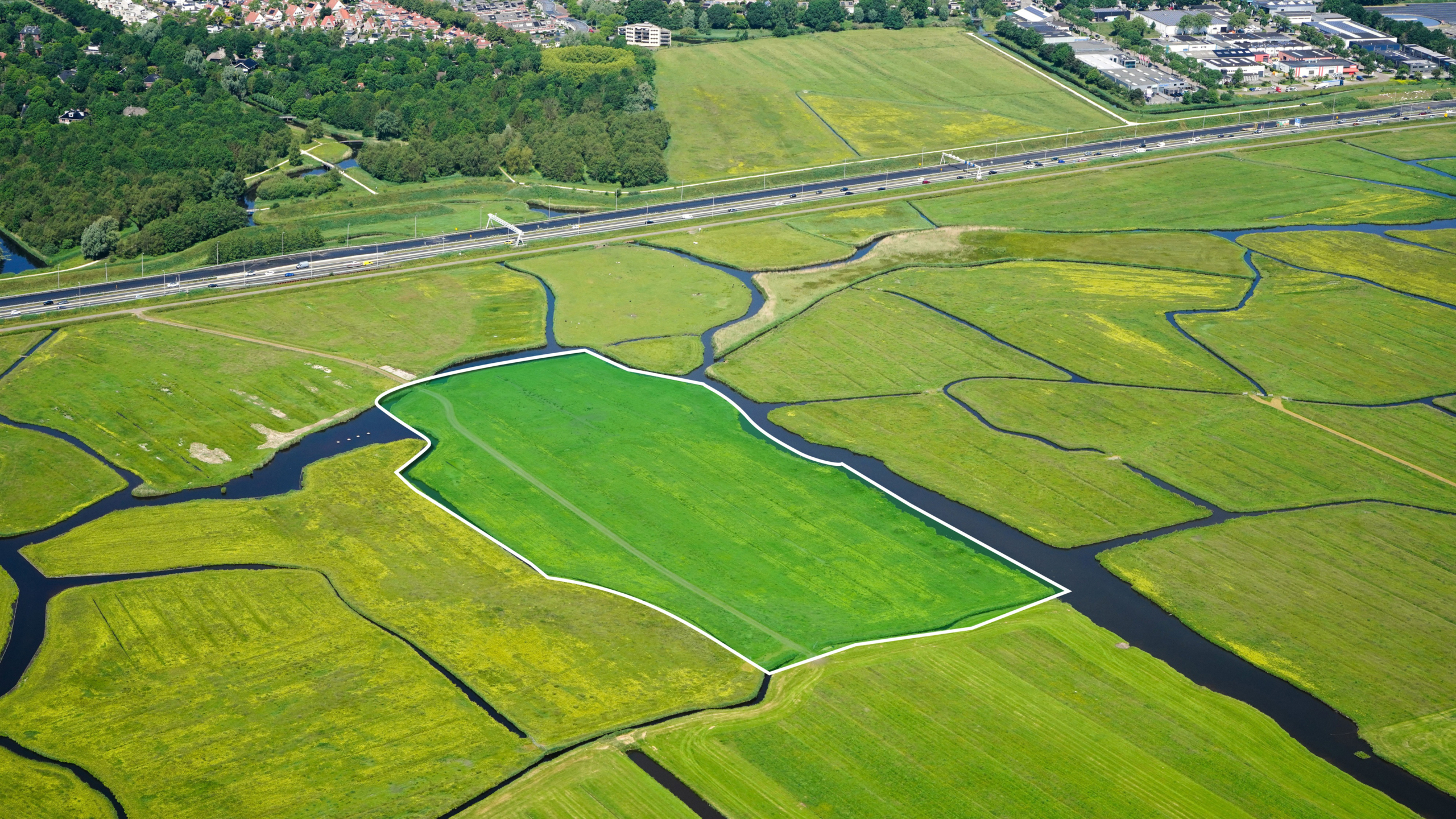 Luchtfoto van polderland met sloten en een groen gemarkeerd perceel met witte omranding naast een snelweg