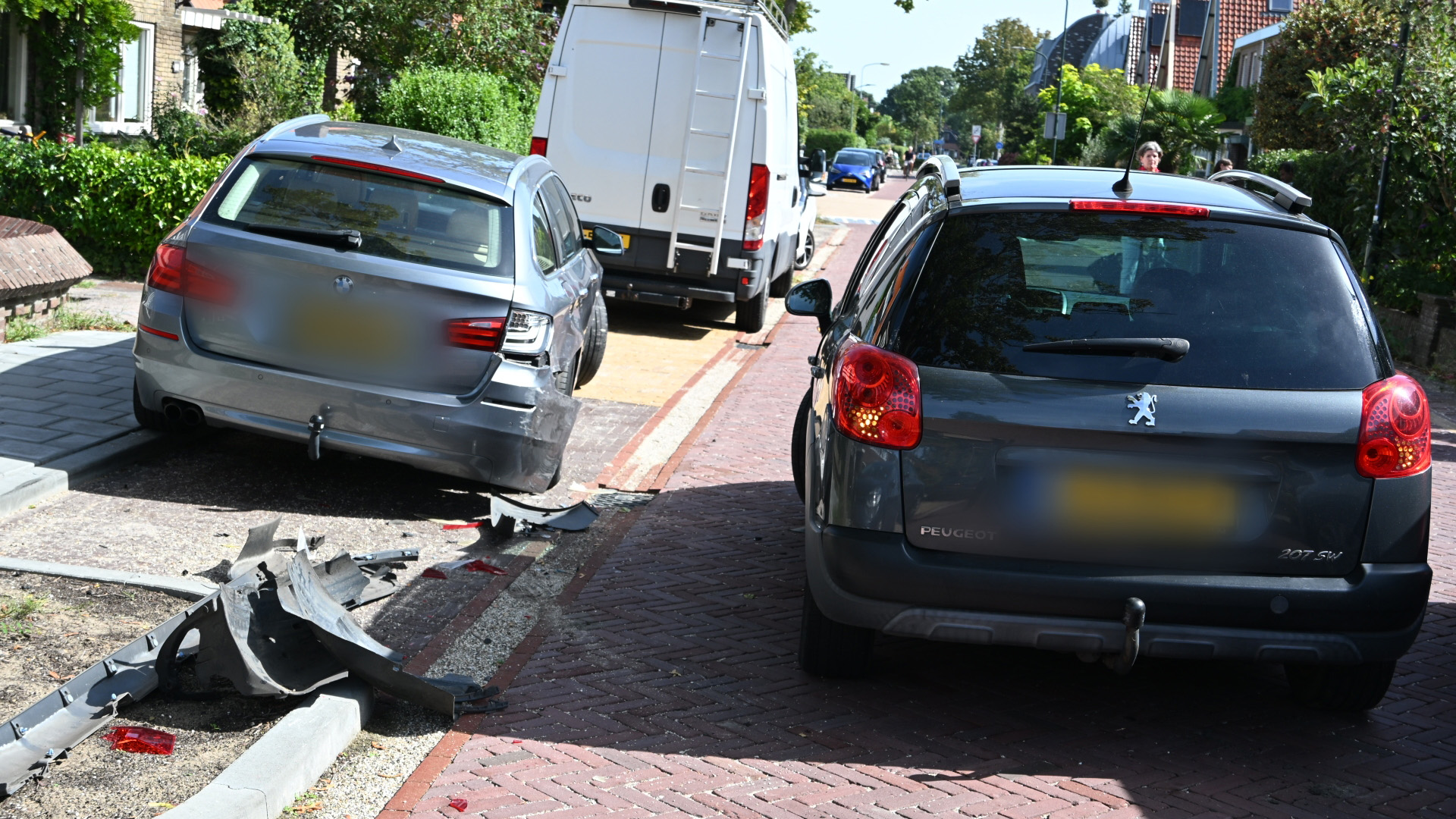 Beschadigde auto na aanrijding met brokstukken op straat, met een bestelbus en geparkeerde auto’s op de achtergrond