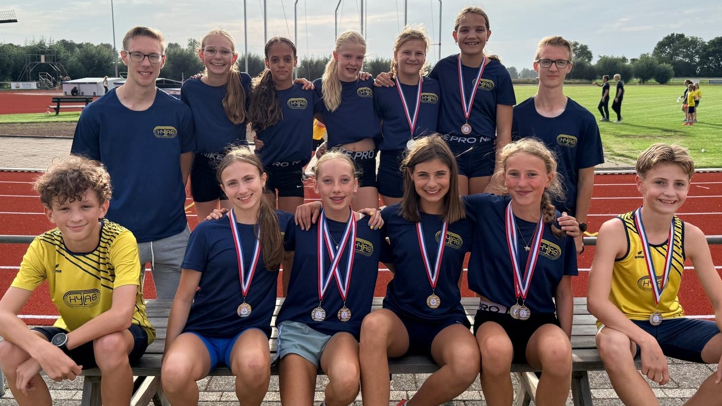 Groep jonge atleten met medailles poseert bij de atletiekbaan, met twee trainers en een sportveld op de achtergrond
