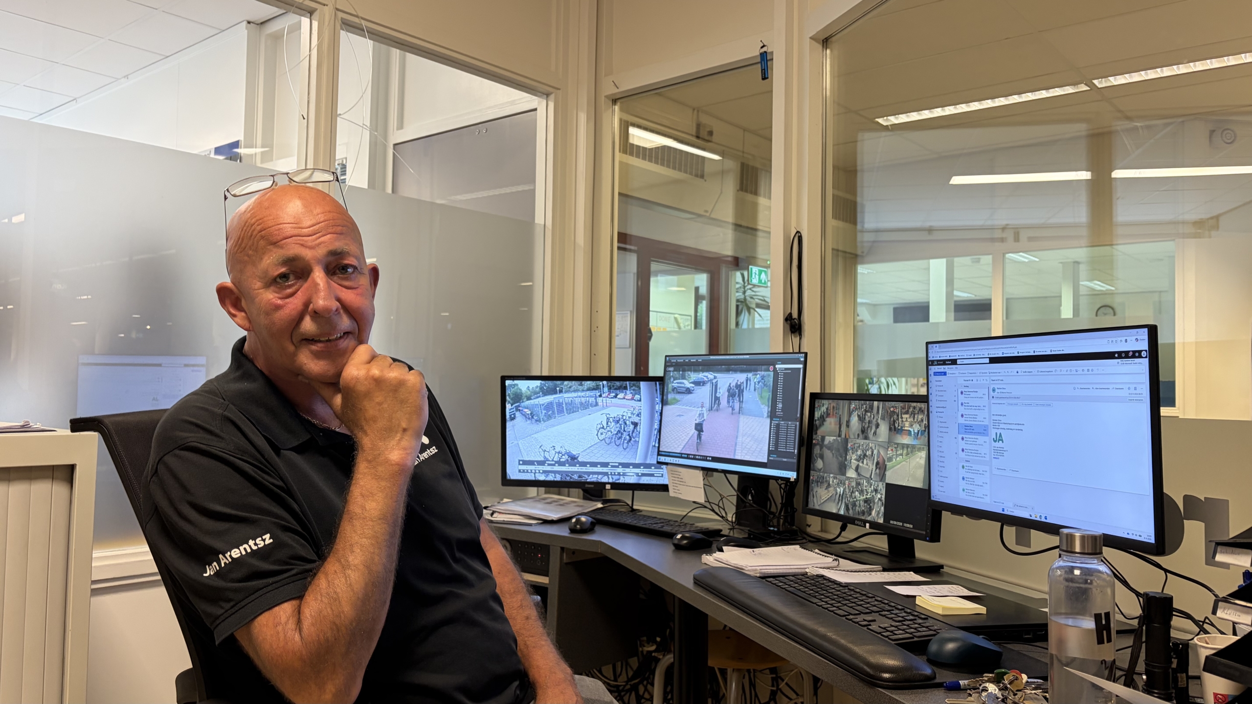 Bewaakte beveiliger zit aan bureau met meerdere monitoren waarop camerabeelden en e-mail op kantoor te zien zijn