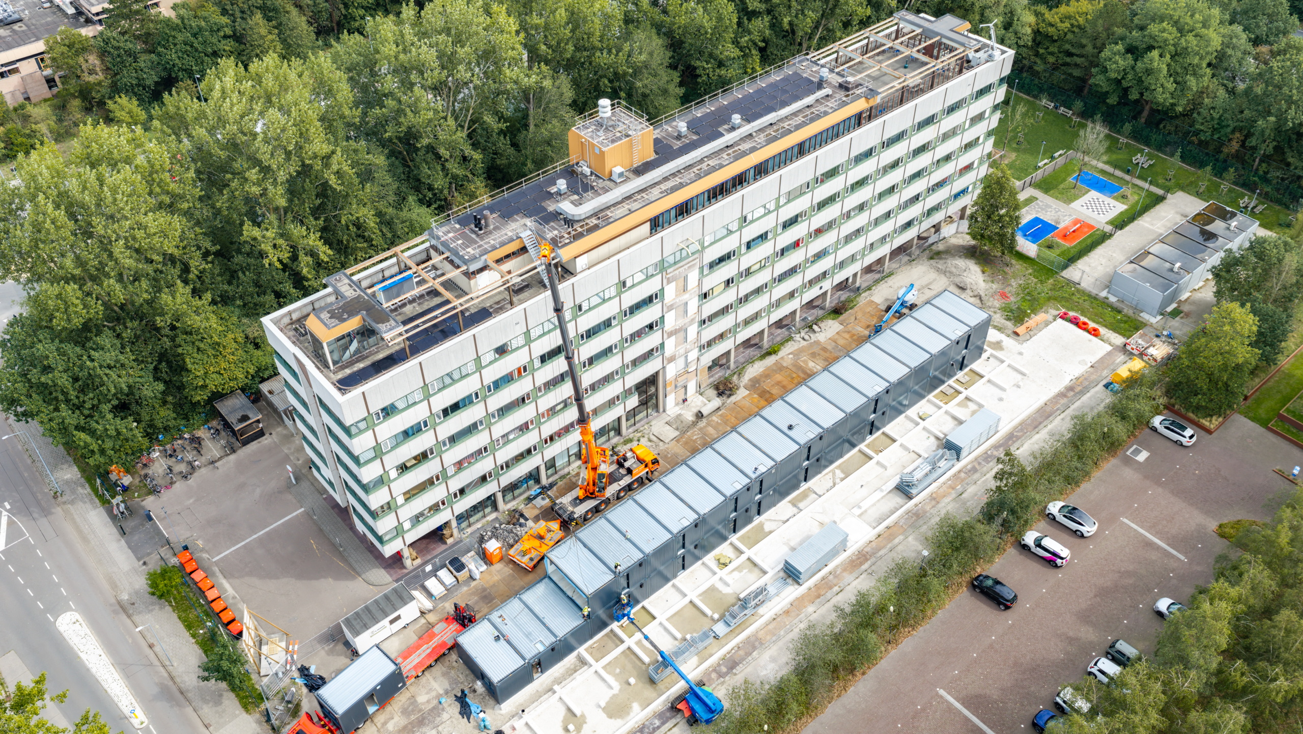 Luchtfoto van een lang flatgebouw met dakrenovatie, kraan en bouwplaats met tijdelijke units naast het gebouw