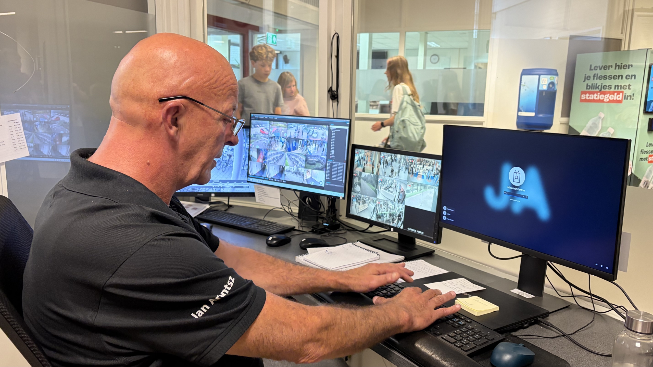 Beveiliger in controlekamer kijkt naar camerabeelden op meerdere monitors, met mensen zichtbaar achter het glas