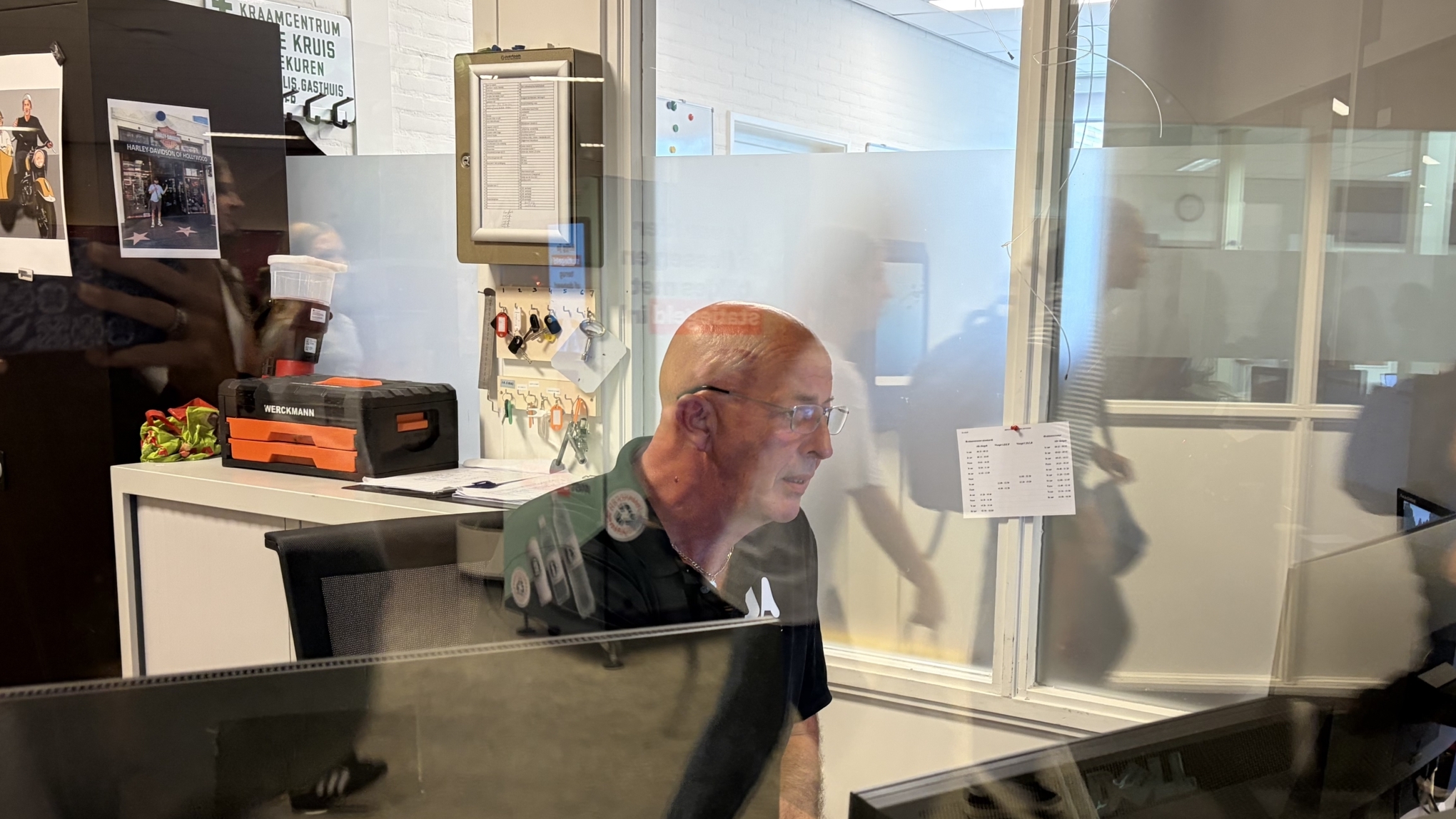 Man met bril zit achter een bureau met monitoren achter glaswand; op de achtergrond lopen mensen langs
