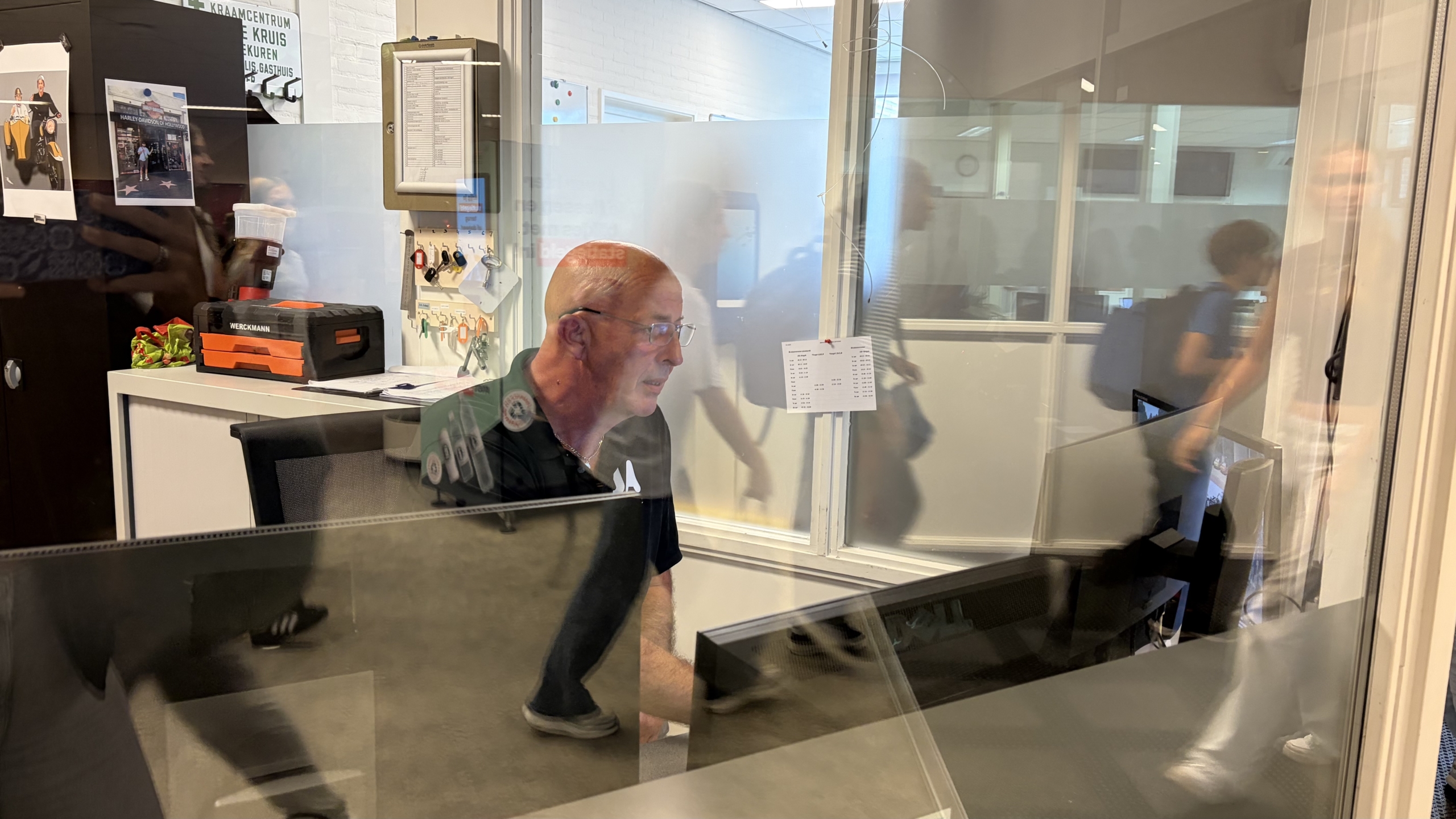 Man met bril zit achter een bureau met monitoren achter glaswand; op de achtergrond lopen mensen langs