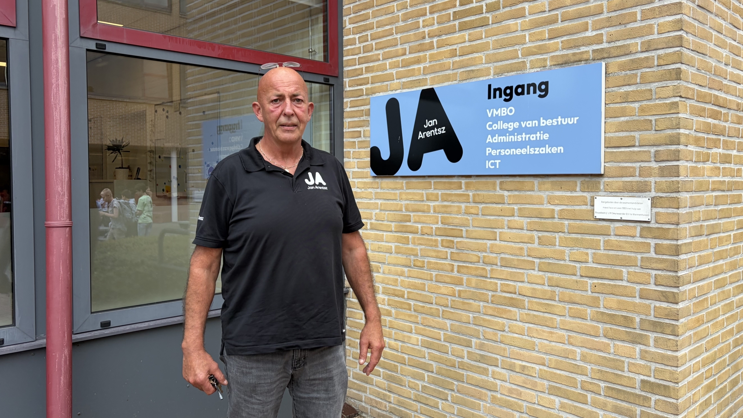 Man in zwart poloshirt bij schoolingang naast bord met tekst Ingang VMBO, College van bestuur, Administratie, Personeelszaken, ICT