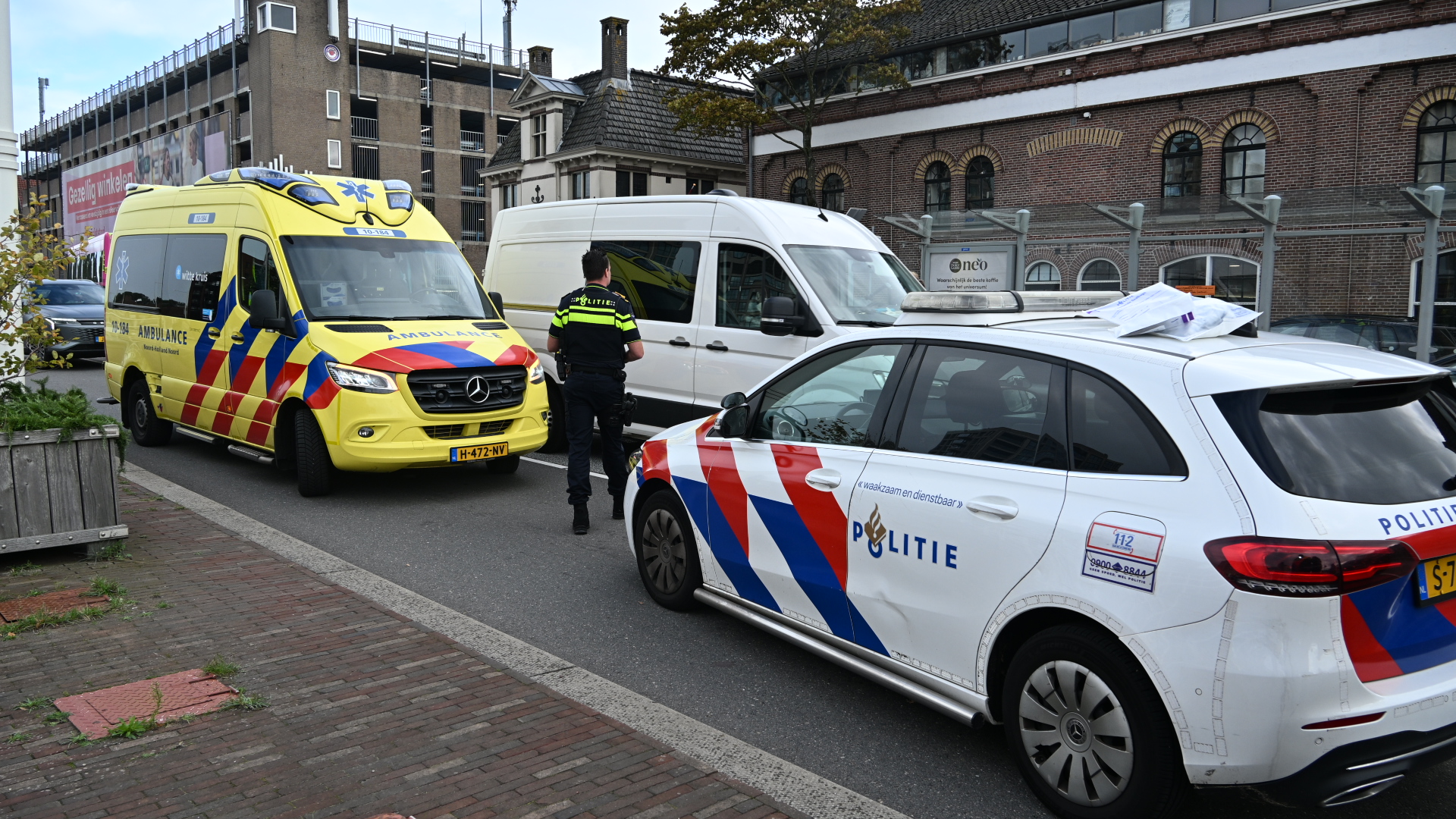 Politieauto en ambulance staan op straat naast een witte bestelbus, met een agent ertussen bij een gebouw