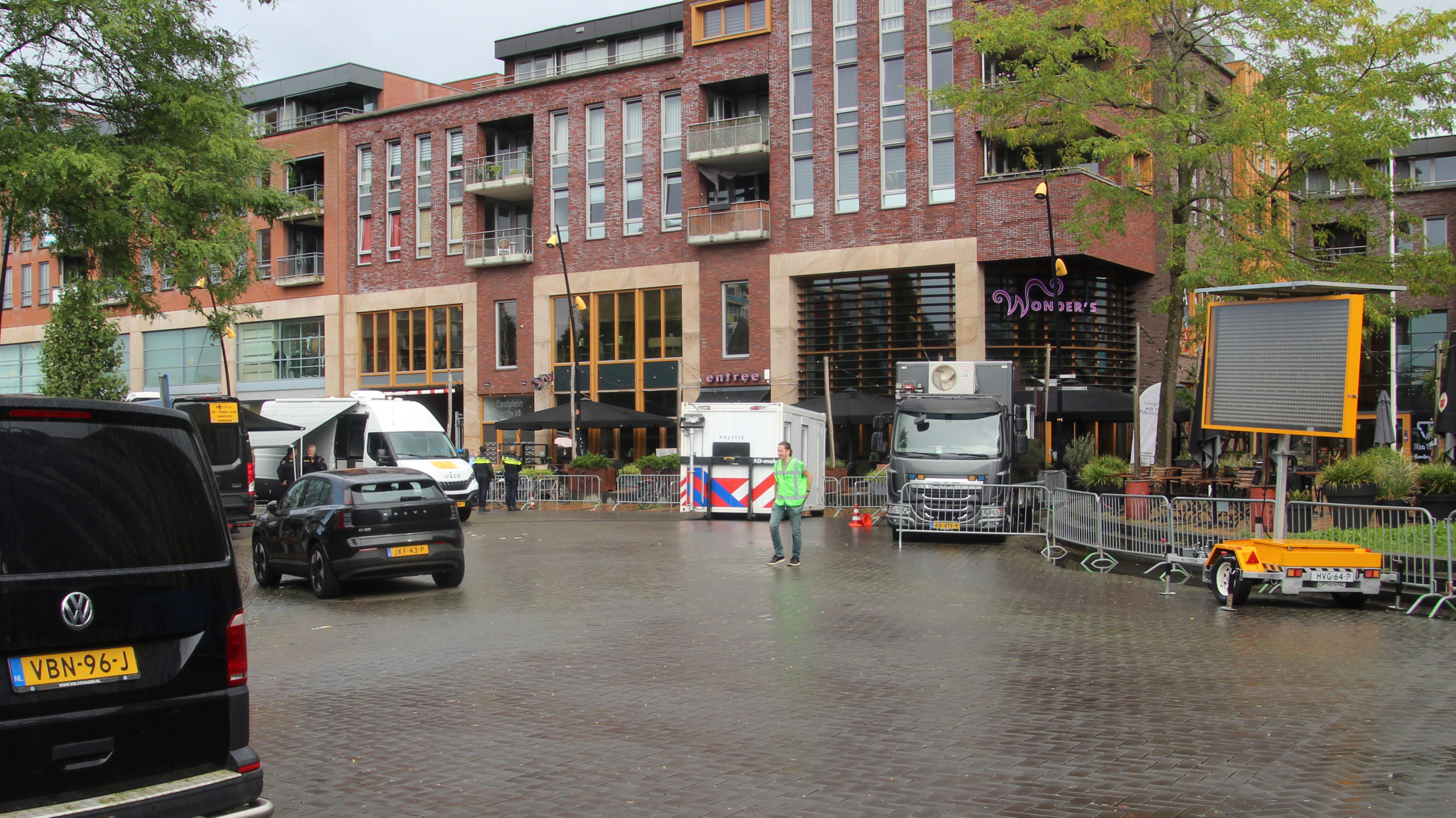 Plein met geparkeerde auto’s en afzethekken voor een bakstenen gebouw met terrassen en het neonbord “Wonder’s”