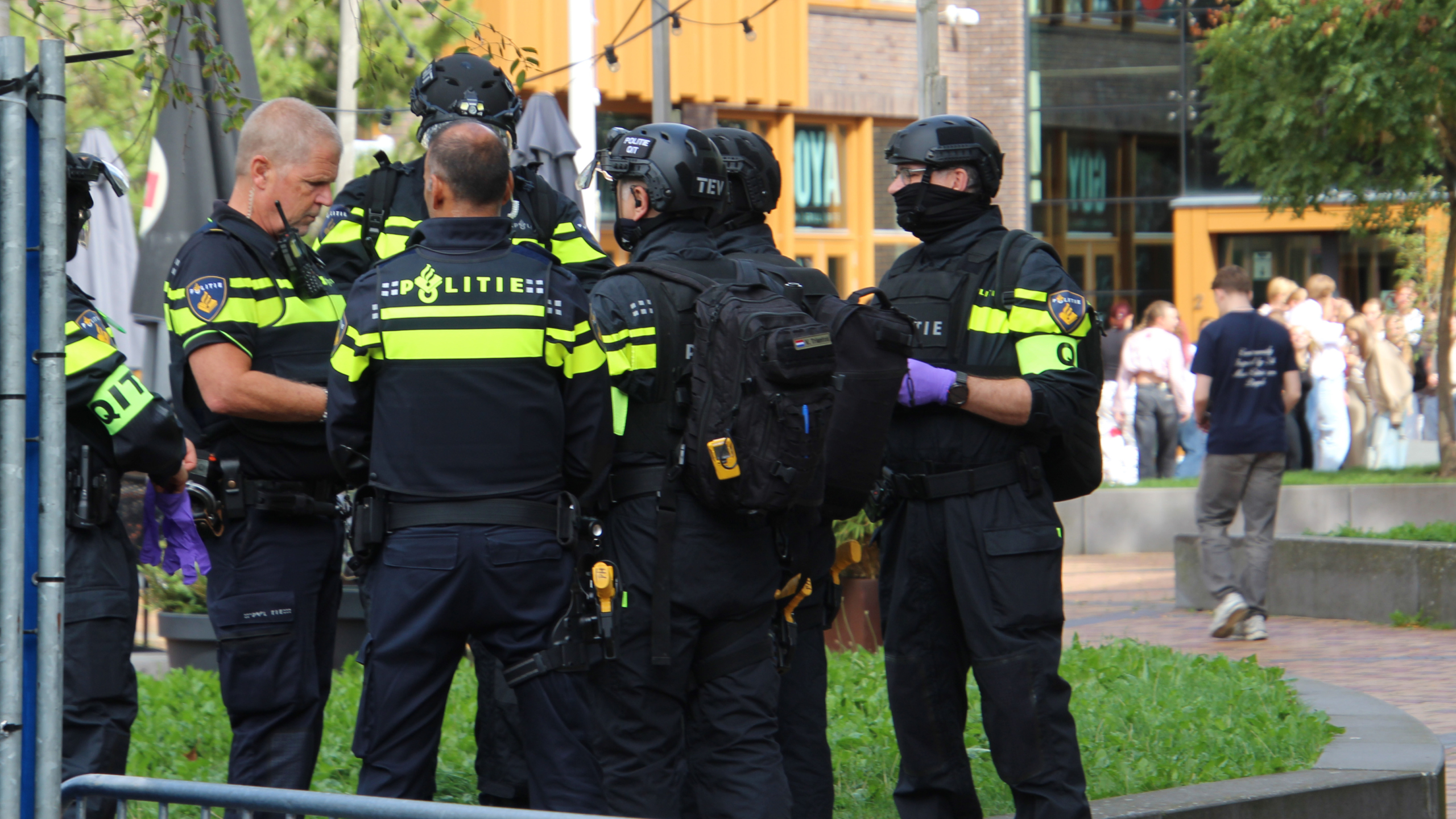 Groep Nederlandse politieagenten in uniform en helmen overlegt op straat, met omstanders op de achtergrond