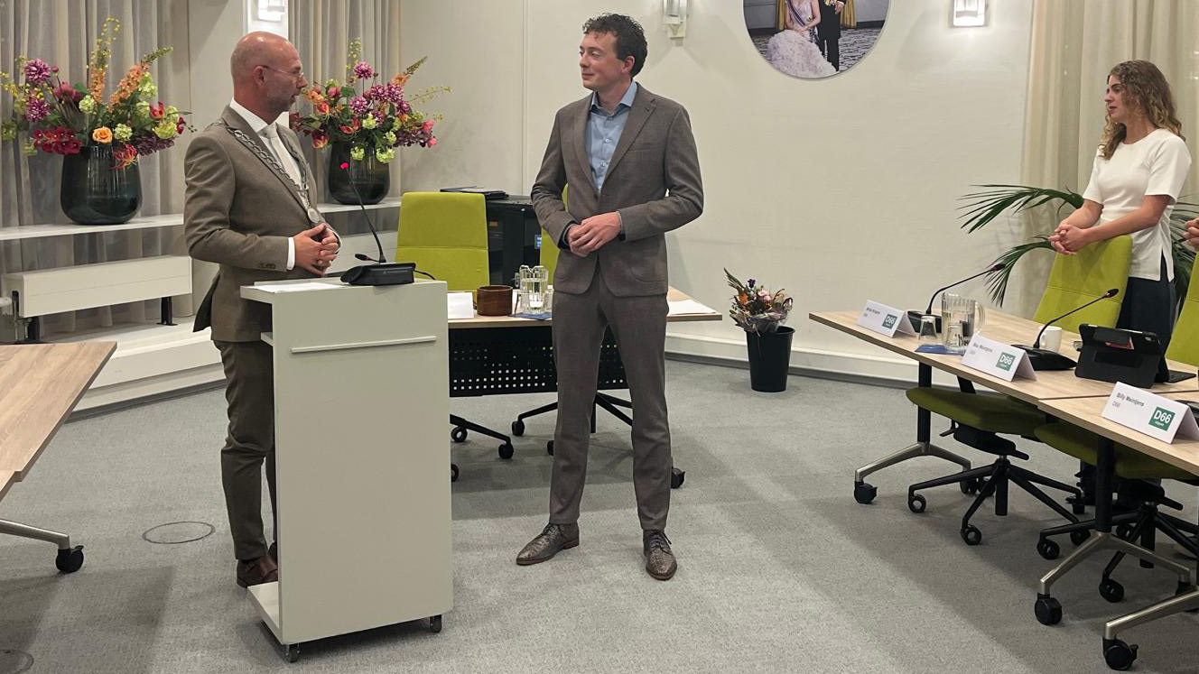 Man met ambtsketen spreekt bij een katheder in raadszaal, terwijl een andere man en vrouw toekijken