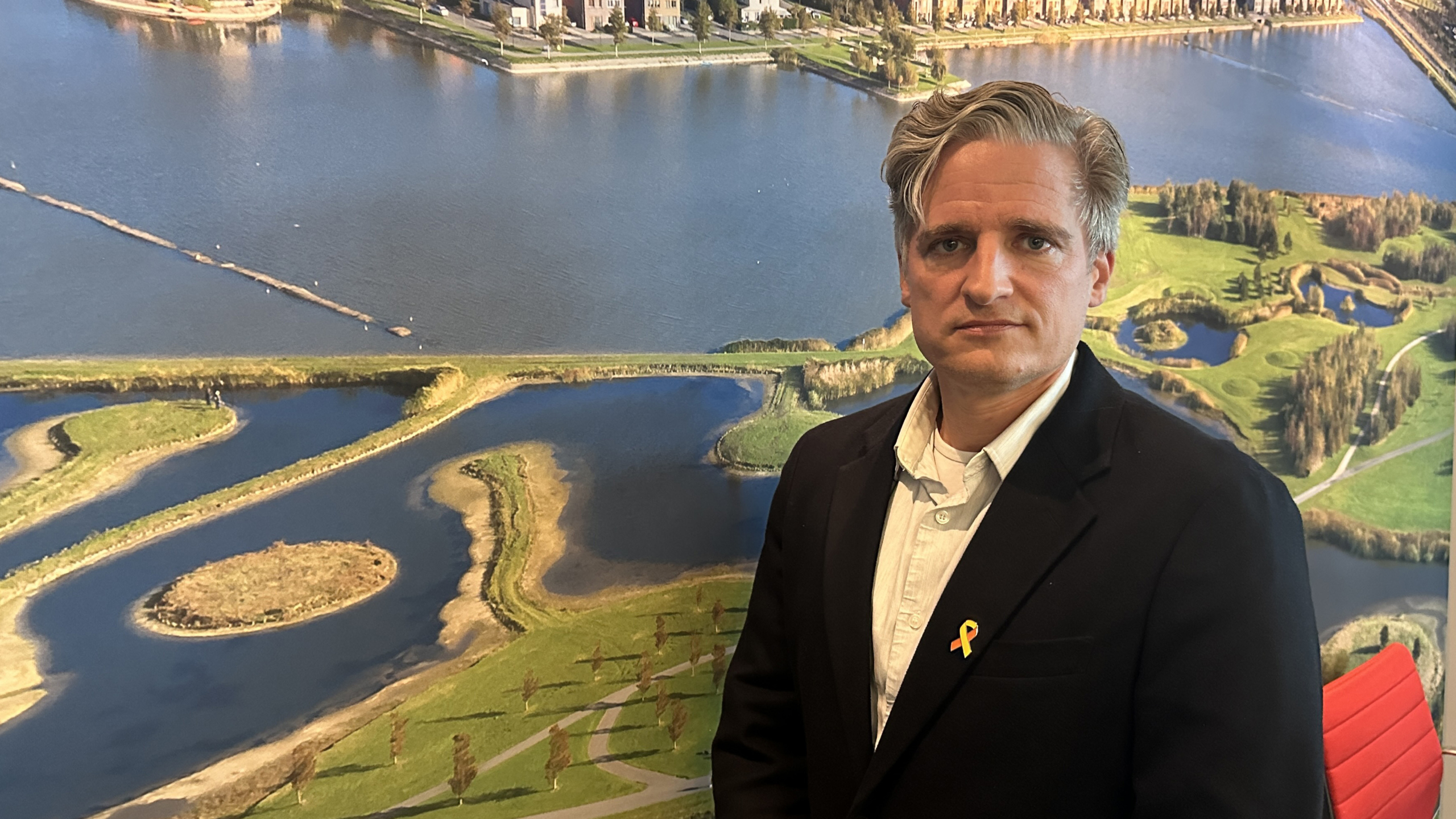 Man in zwart colbert met gekleurde pin, staand voor een luchtfoto van water met eilanden en groen parklandschap